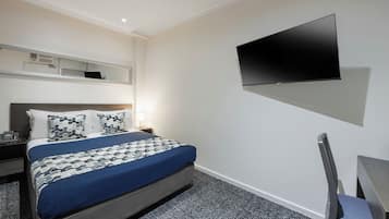 Quarto de hotel com cama, escrivaninha, cadeira, TV de tela plana na parede e uma mesinha.