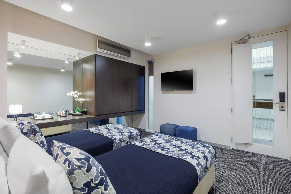 Habitación de hotel con dos camas, un escritorio, un televisor de pantalla plana, un espejo y una puerta con panel de vidrio.