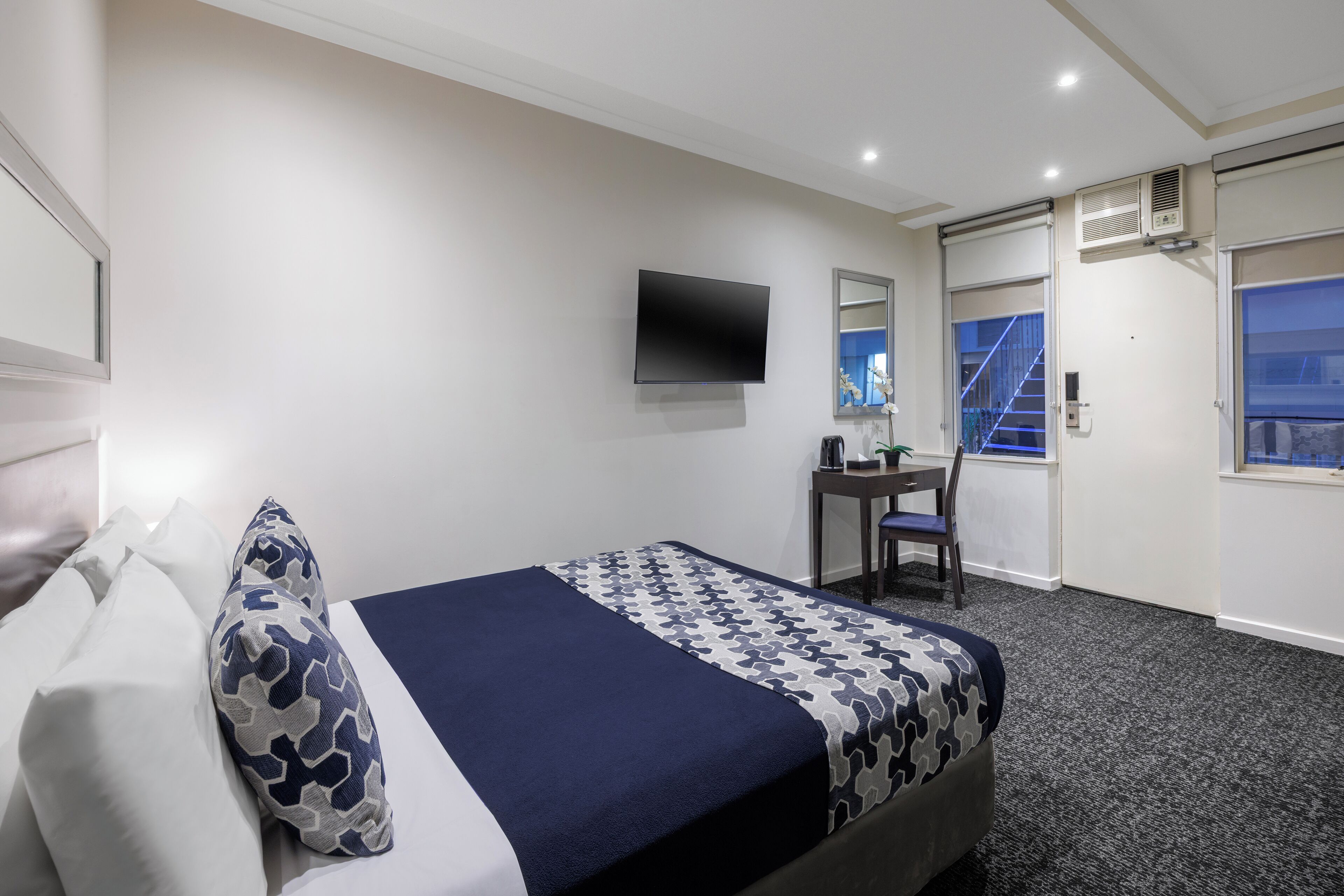 A hotel room with a bed, a desk, a chair, a TV, and a window with a view of a staircase.