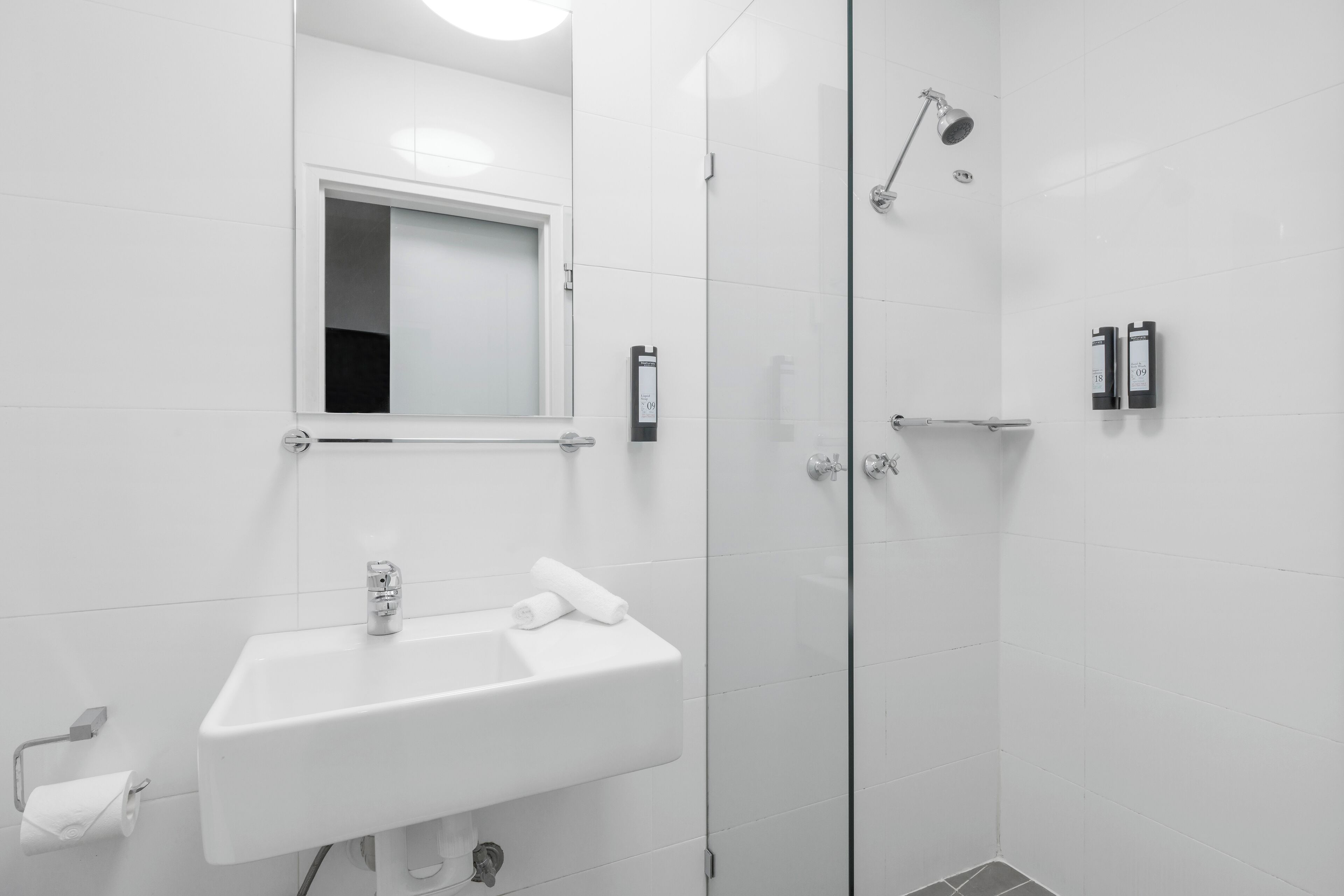 A modern bathroom with a white sink, a mirror, a shower with a glass door, and two towel dispensers.