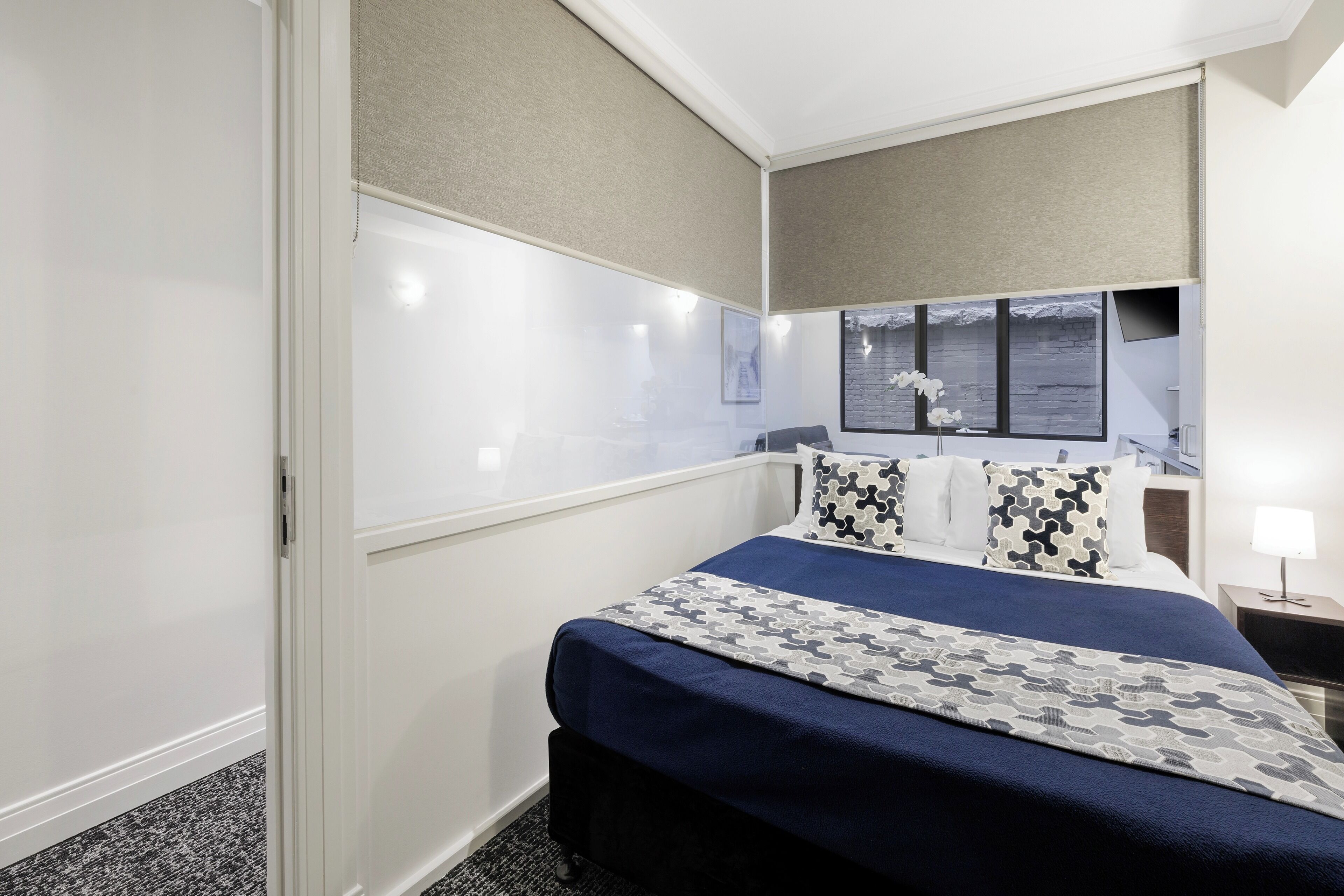 A hotel room with a large bed, a nightstand with a lamp, a window with a view, and a wall-mounted shelf.