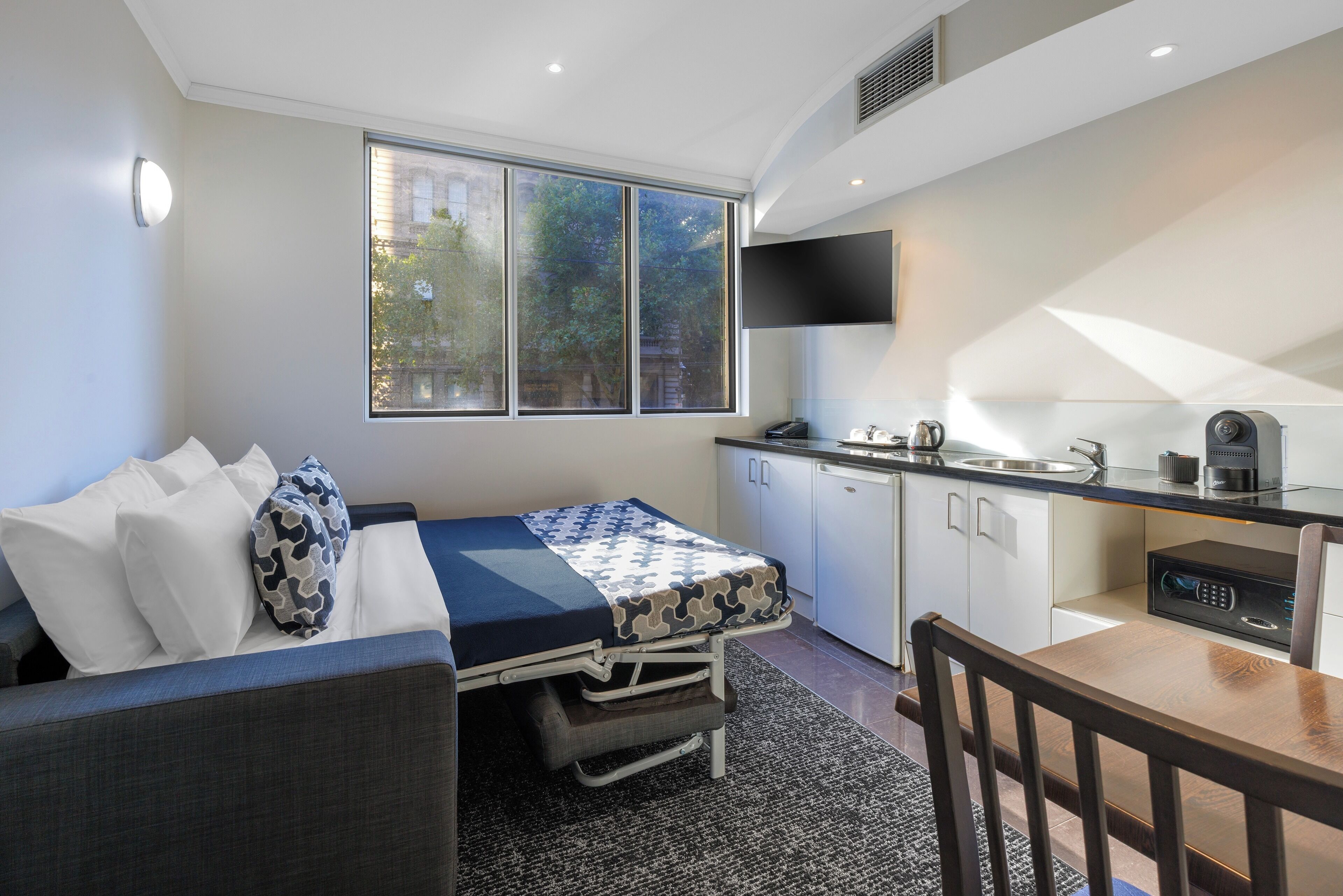 Una habitación compacta con cama, una kitchenette pequeña, una mesa de comedor y televisor instalado en la pared.