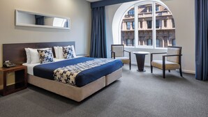 Quarto de hotel com uma cama grande, uma escrivaninha, uma cadeira, uma mesa redonda e vista para um edifício através de uma janela ampla.