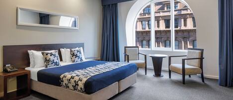 Habitación de hotel con una cama grande, un escritorio, una silla, una mesa redonda y vistas a un edificio a través de un ventanal.
