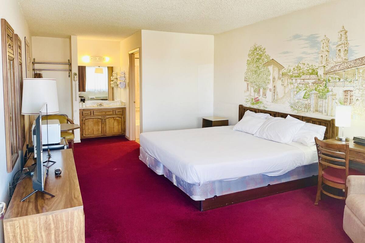 A hotel room with a large bed, a wooden desk, a desk lamp, a chair, a bedside table, a mirror, a bathroom with a sink and towel rack, and a mural on the wall.