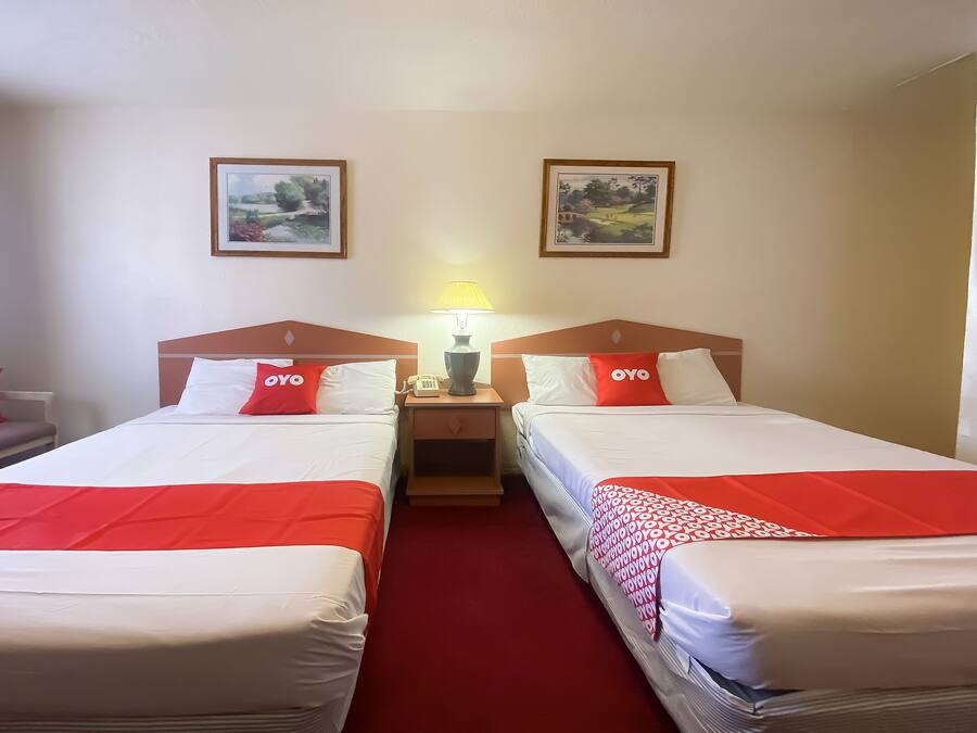 Habitación de hotel con dos camas, cada una con una colcha rojo y blanco, una mesita de noche con lámpara, y dos cuadros enmarcados en la pared.