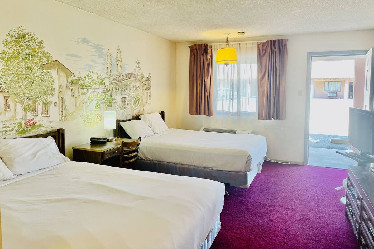 A hotel room with two beds, a desk with a lamp, a mural on the wall, and a view of the outside.