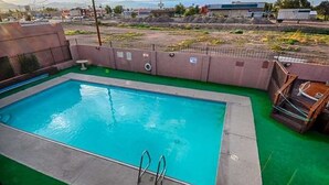 A rectangular pool with a green deck, surrounded by a fence, and a view of a cityscape in the background.
