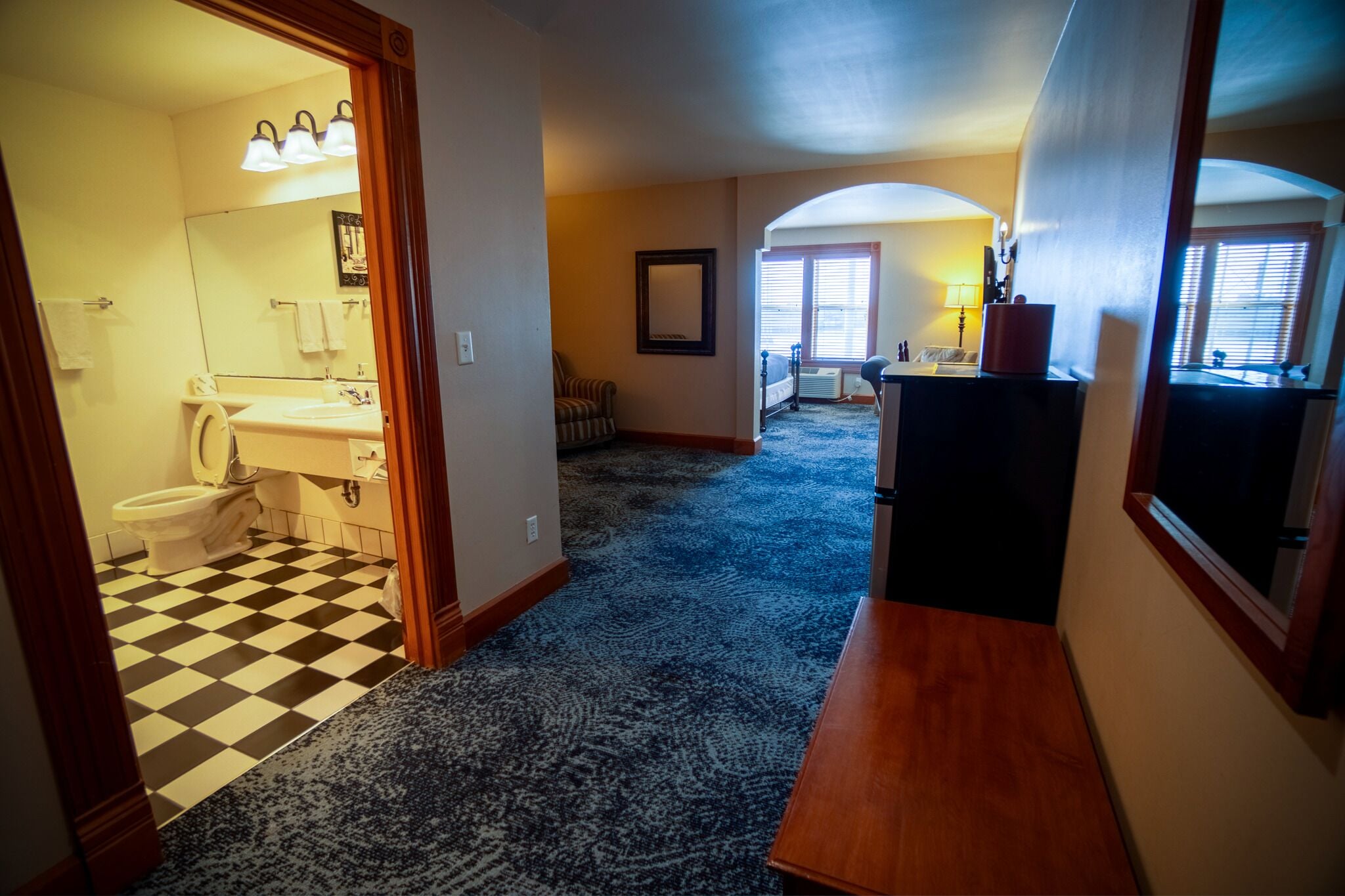 A hallway with a checkered floor leading to a bathroom with a sink and toilet. There is a chair and a lamp in the room.