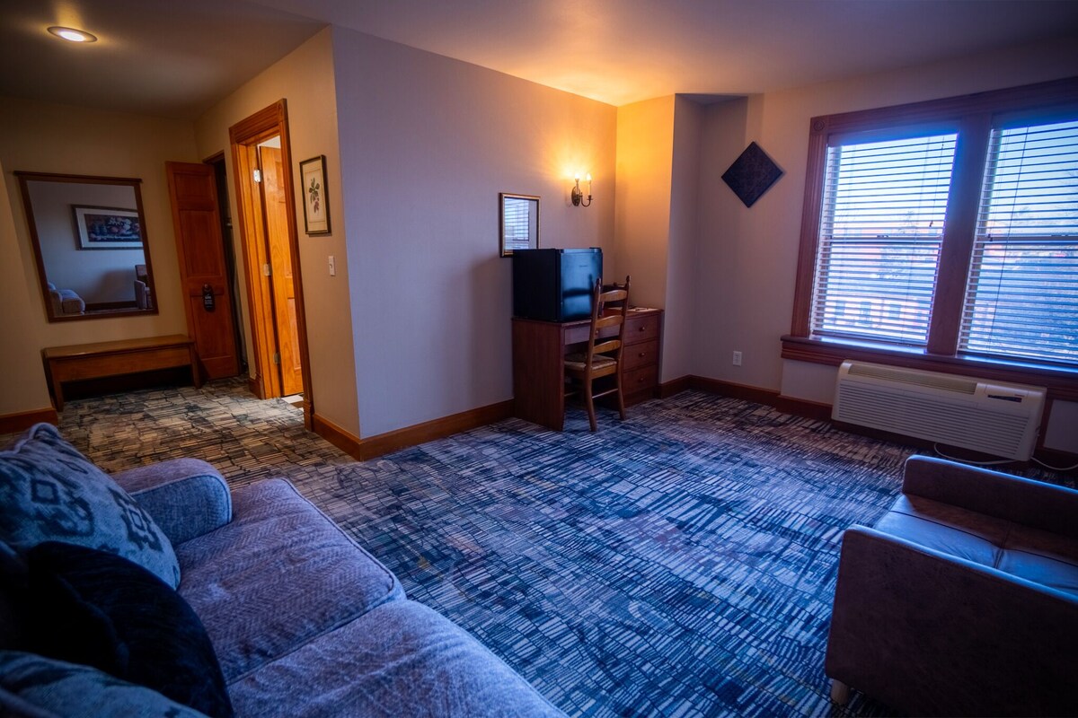 A living room with a blue patterned carpet, a blue sofa, a wooden desk with a chair, and a television.
