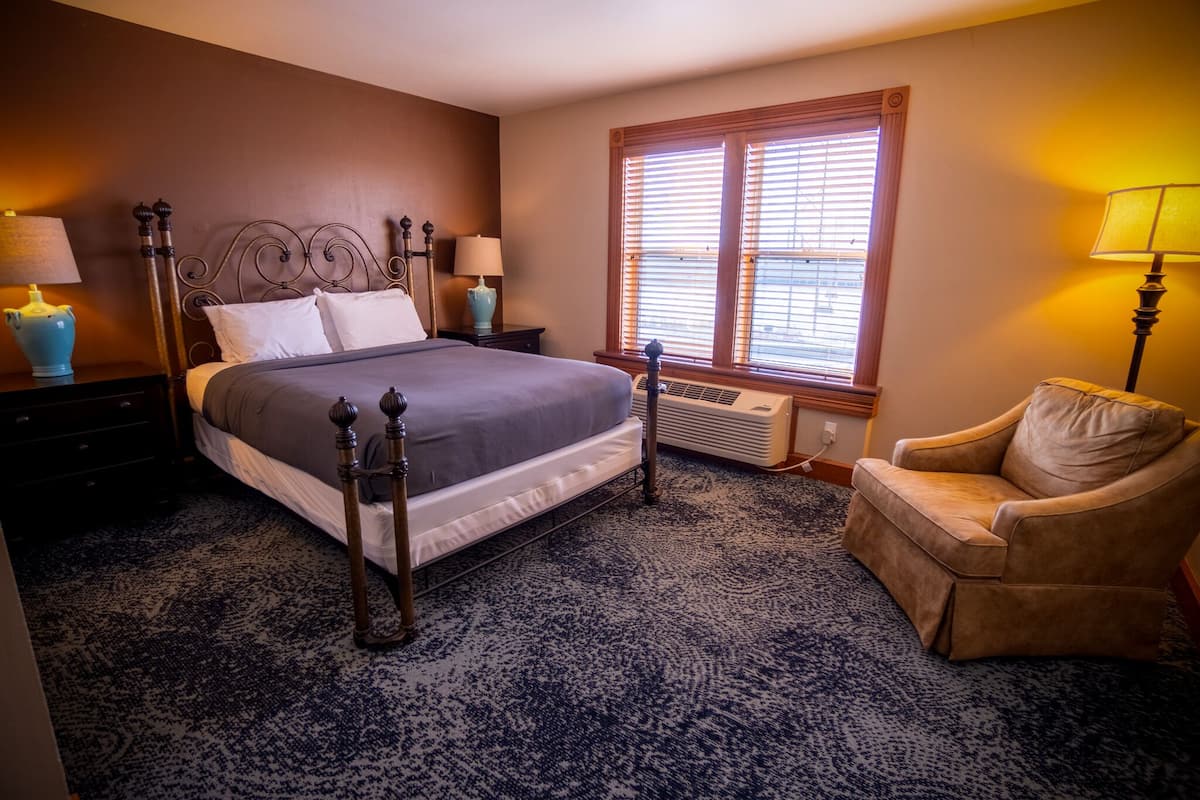 A bedroom with a large bed, two bedside tables with lamps, a chair, and a window with blinds.