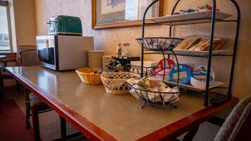 A dining area with a wooden table, wicker baskets, a metal shelf with food items, a microwave, and a refrigerator.