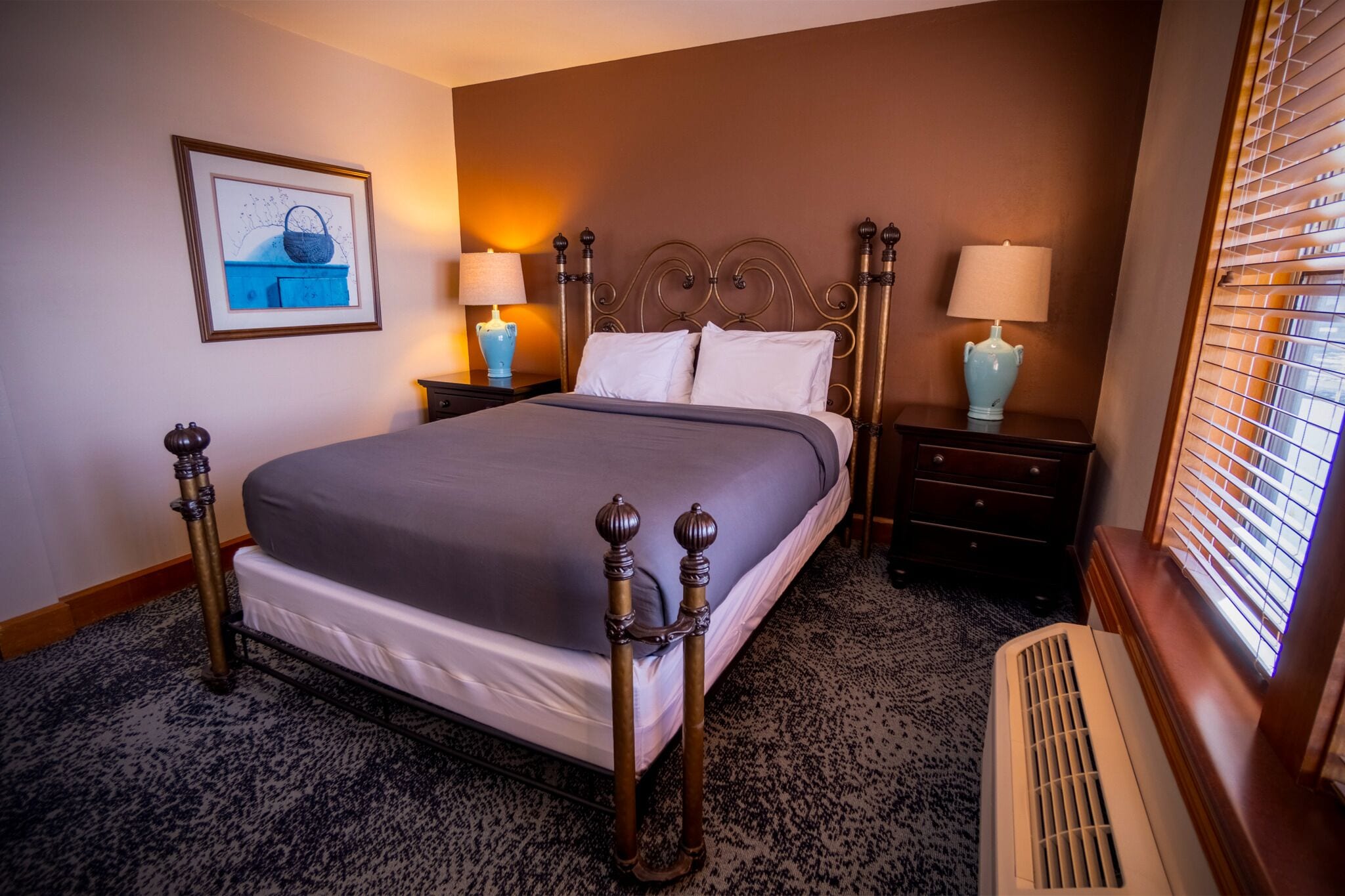 Una habitación de hotel con una cama grande, dos mesitas de noche con lámparas, un cuadro enmarcado en la pared y una ventana con persianas.