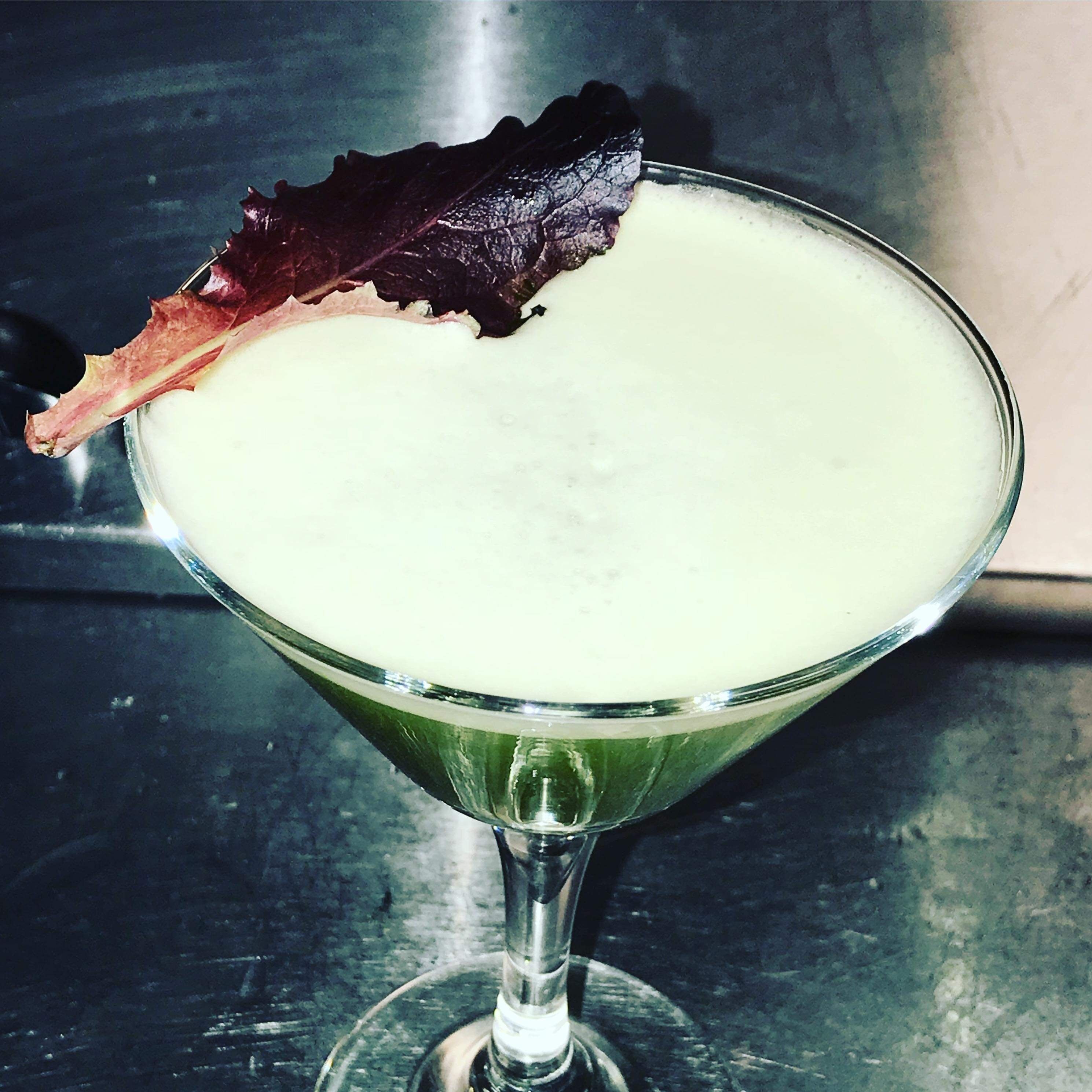 A martini glass with a green liquid, garnished with a leaf and a slice of red fruit.