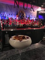 Un bar con varias botellas y una copa de martini con decoración de postre sobre una encimera de mármol.