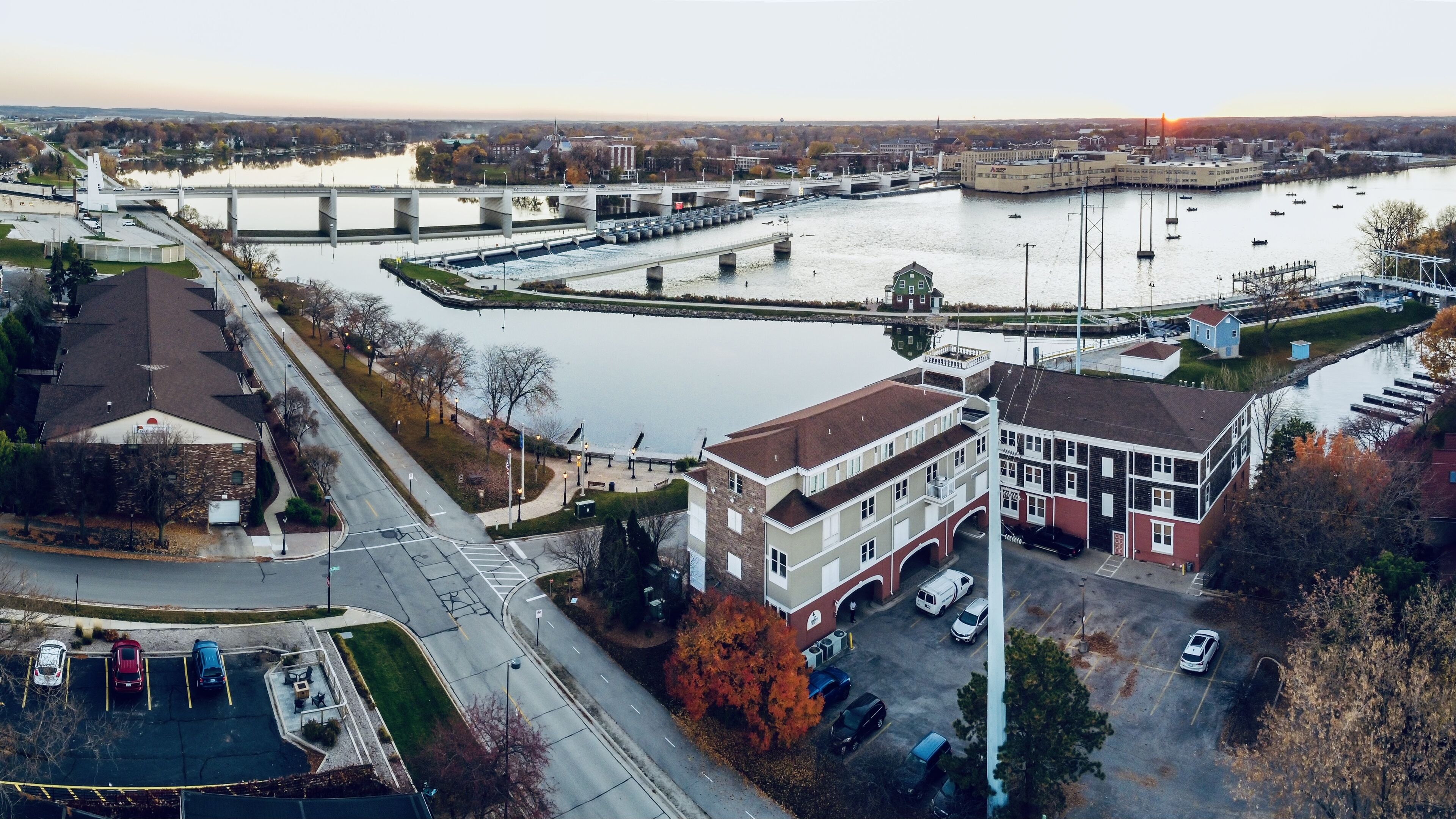An aerial view of a riverside area with a bridge, buildings, and a parking lot.