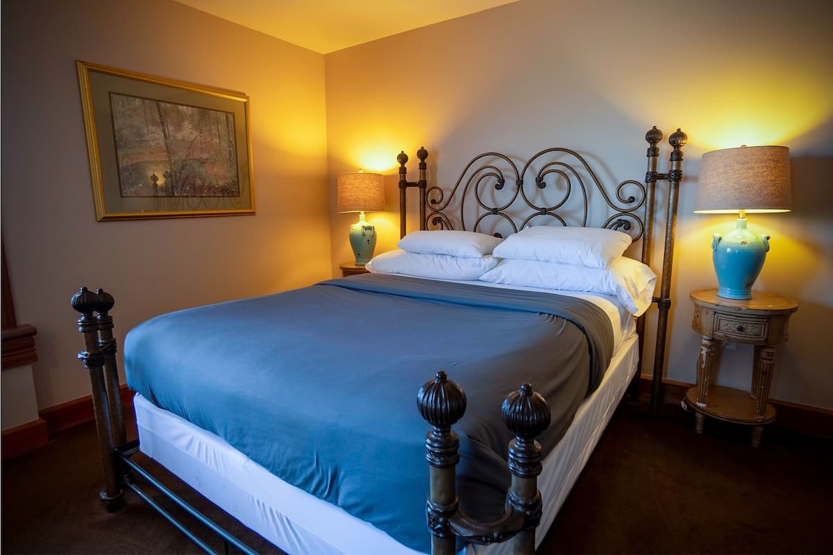 A neatly made bed with a blue bedspread and white pillows, flanked by two bedside tables with lamps, in a room with a framed artwork on the wall.