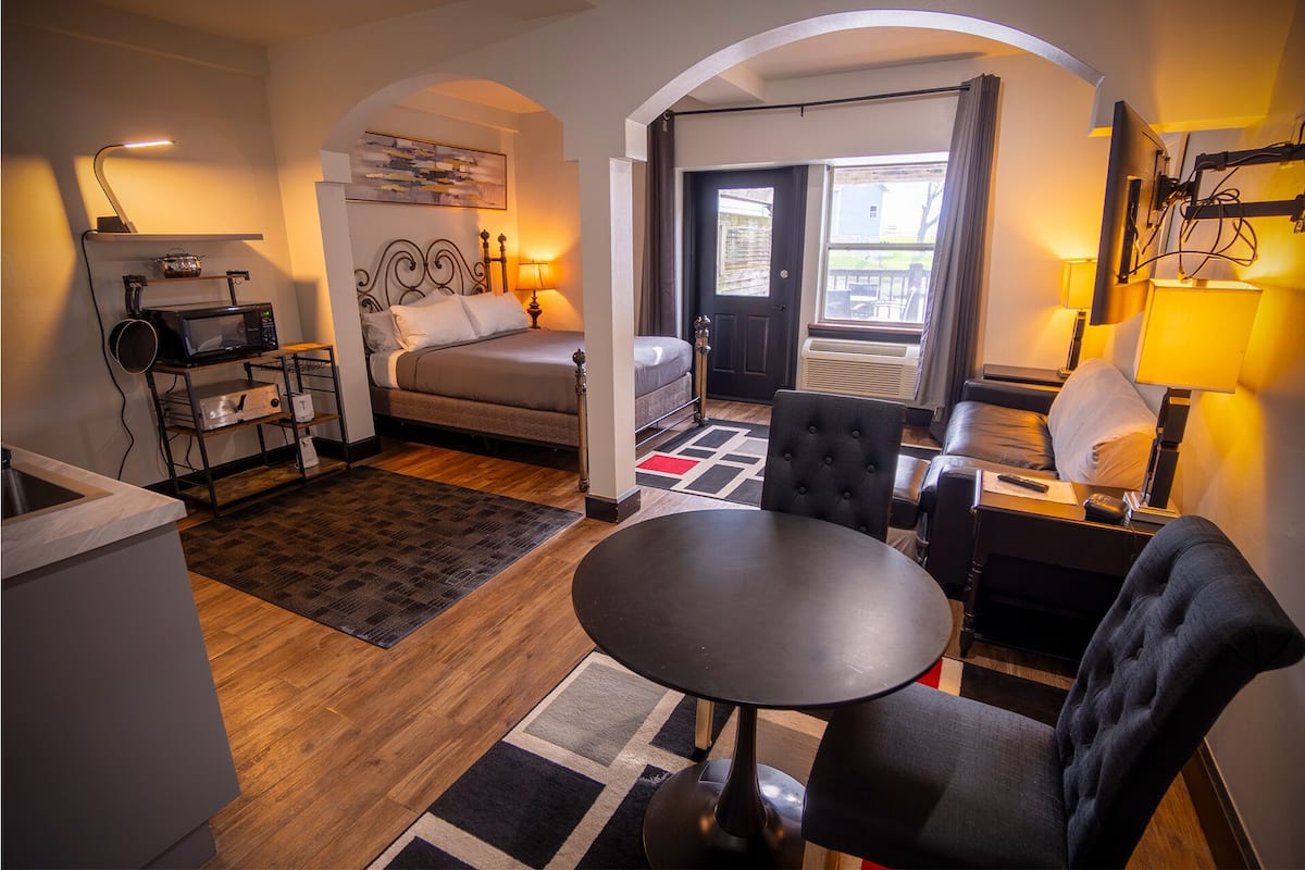 A hotel room with a bed, a dining table, a sofa, a kitchen area with a microwave, and a view of the outside.