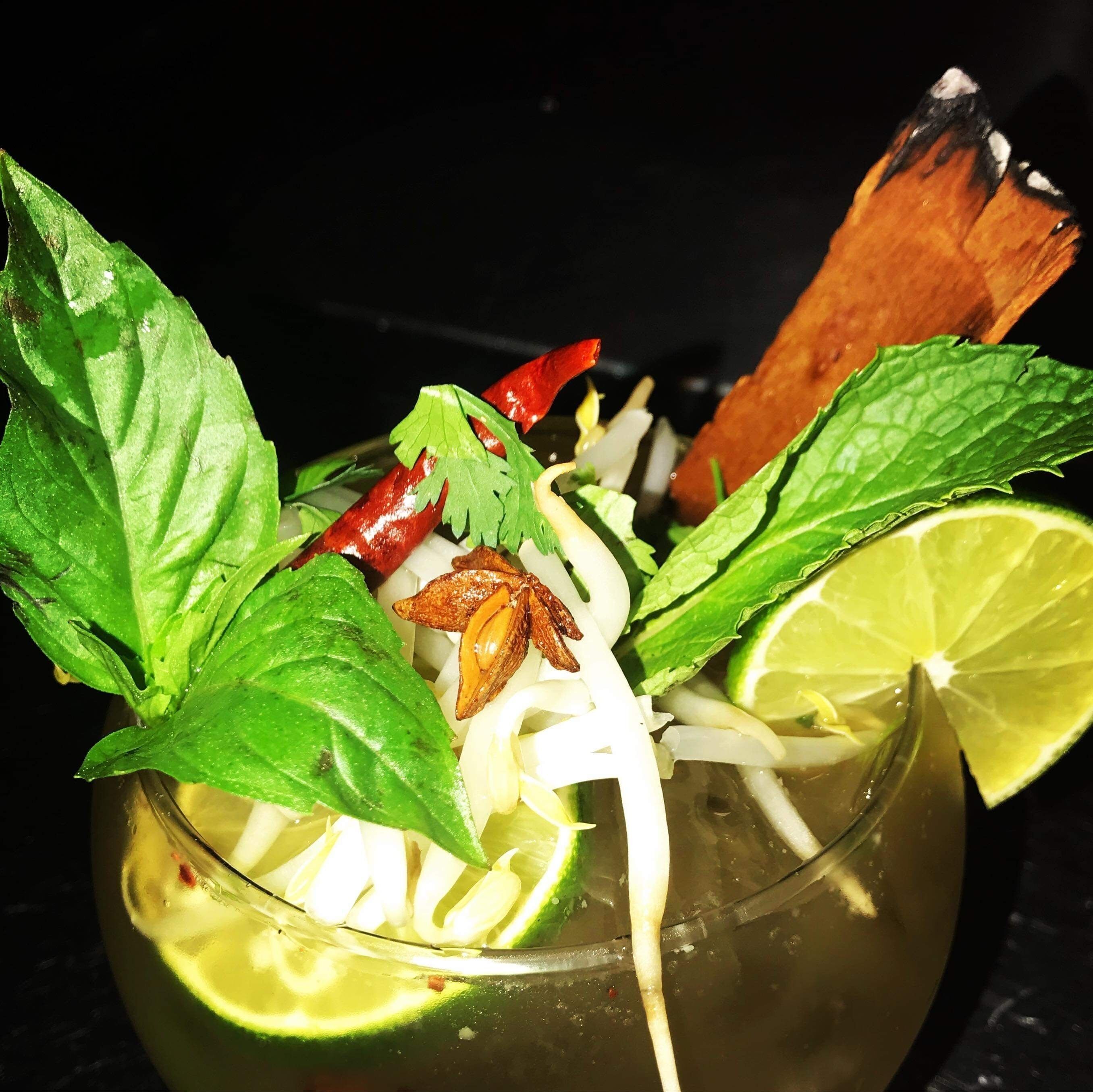 A glass containing a beverage with lime slices, mint leaves, red chili, and star anise.