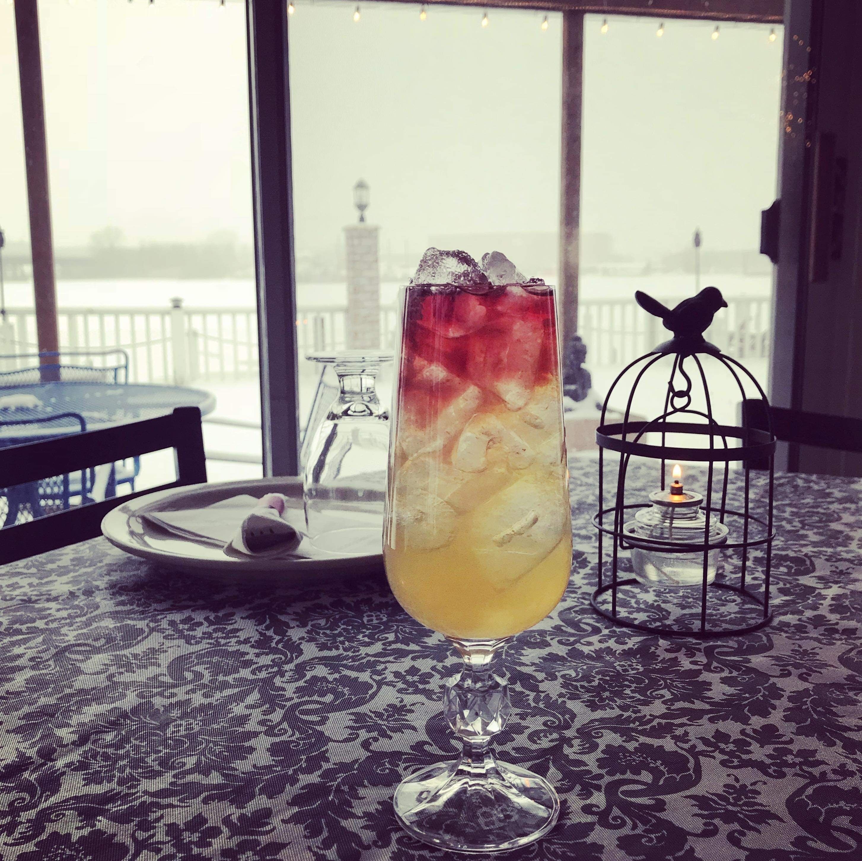 Un verre rempli d’une boisson glacée est posé sur une table, à côté d’un chandelier en forme de cage à oiseaux et d’une assiette avec une serviette. De grandes fenêtres offrent une vue sur une étendue d’eau.