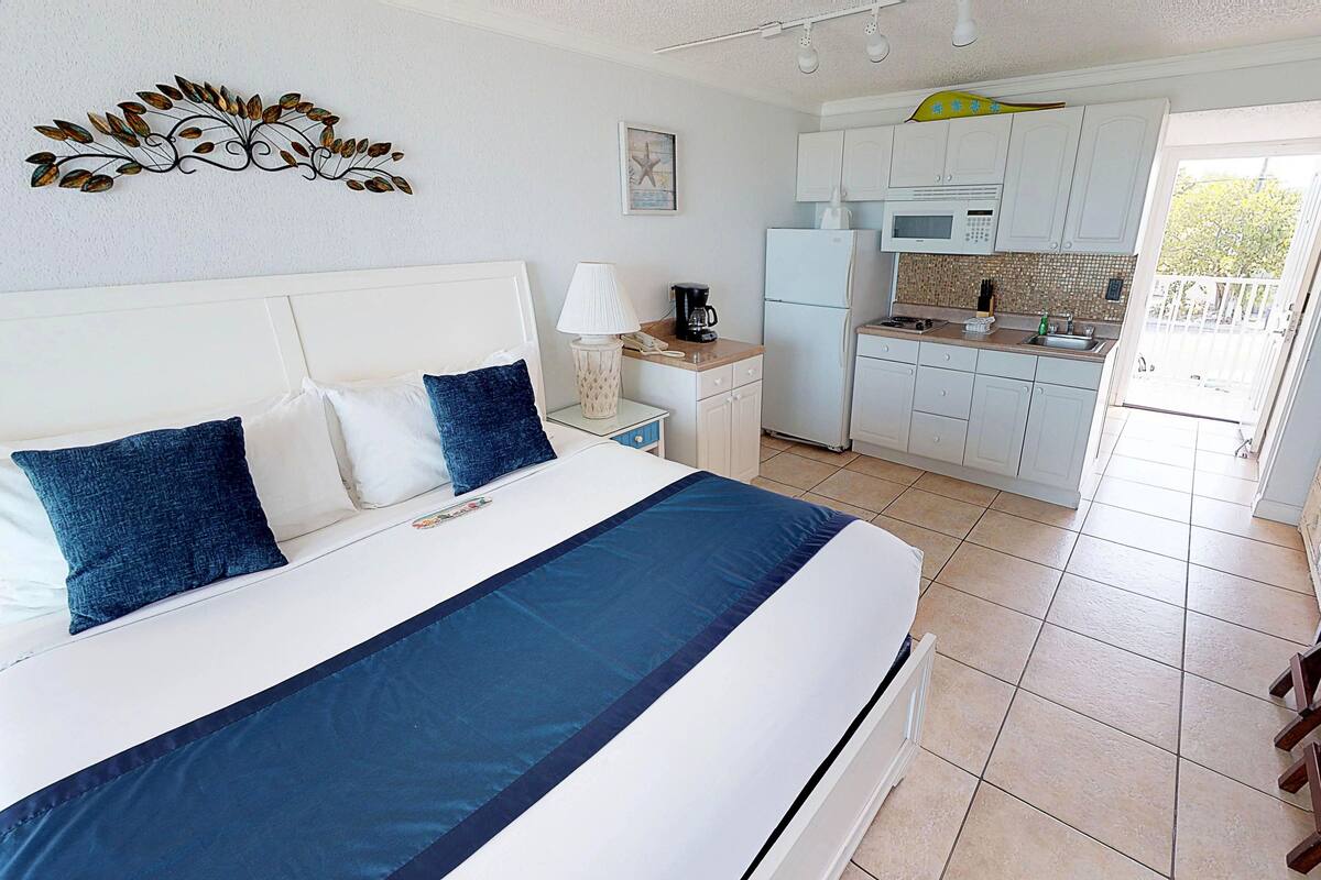 A bedroom with a large bed, a nightstand, a coffee maker, and a view of the kitchen.
