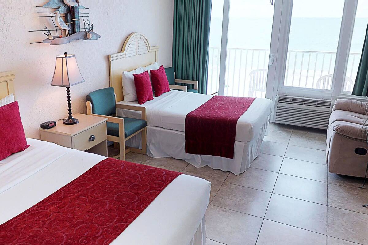 A hotel room with two beds, a chair, a nightstand, a lamp, and a view of the sea.