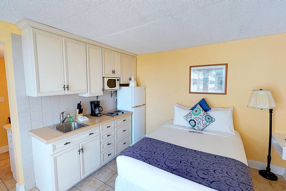 A compact hotel room with a kitchenette, a bed with patterned bedding, a bedside lamp, and a framed picture on the wall.