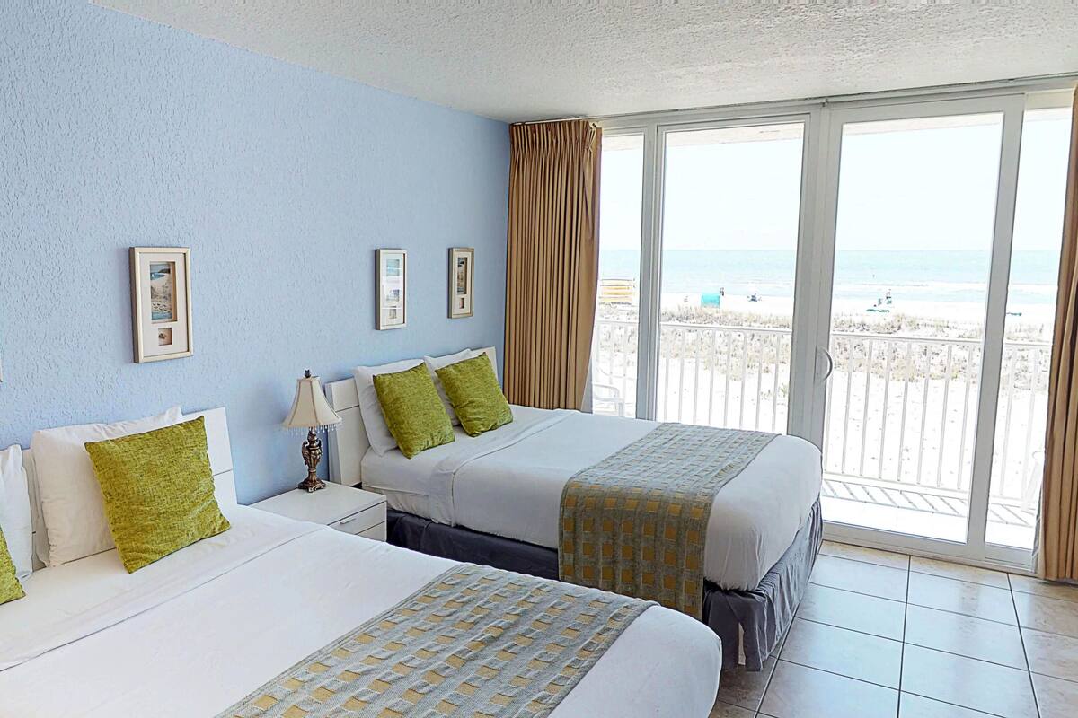 A hotel room with two beds, a balcony with a view of the beach, and framed pictures on the wall.