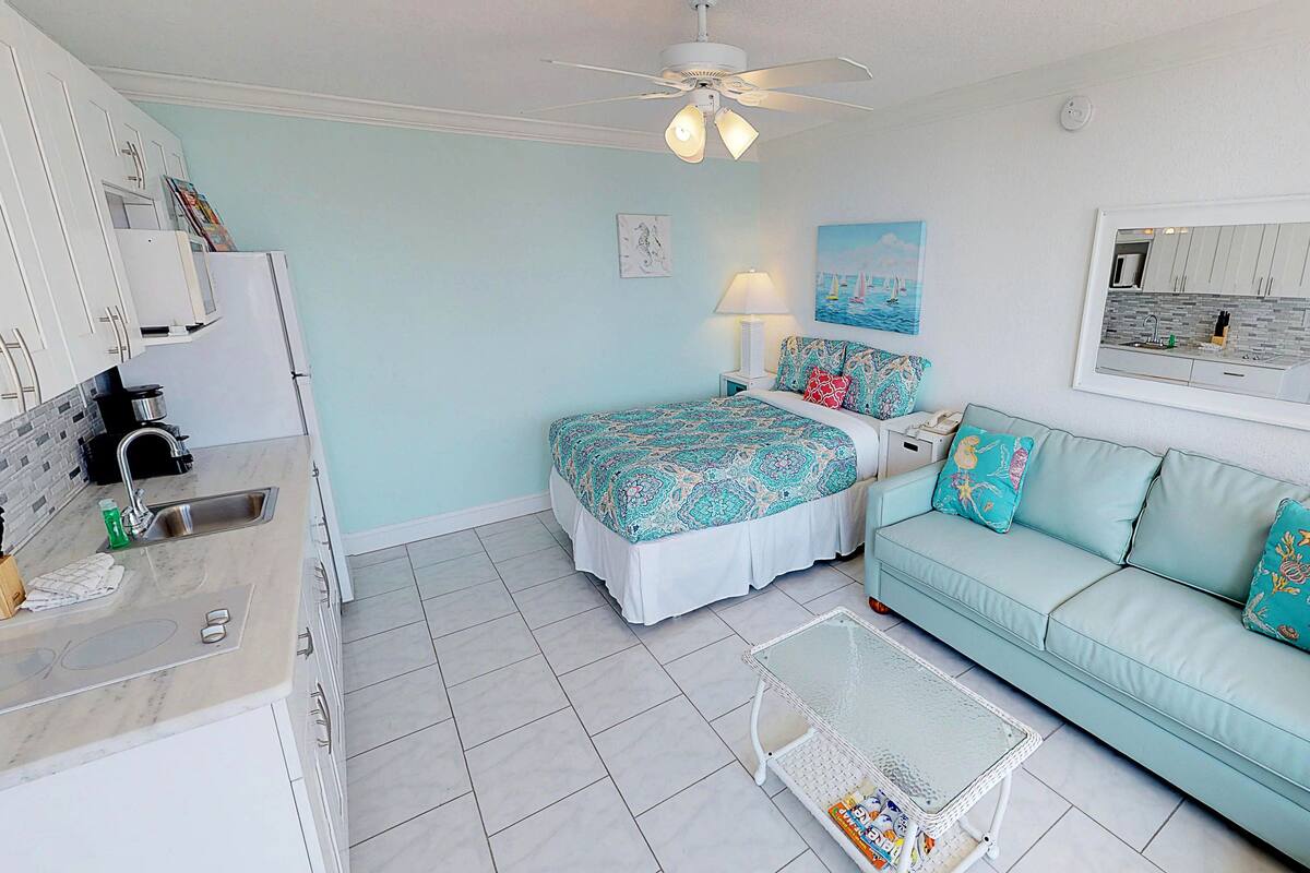 A compact hotel room with a kitchenette, a bed with patterned bedding, a light blue sofa with decorative pillows, and a small table.