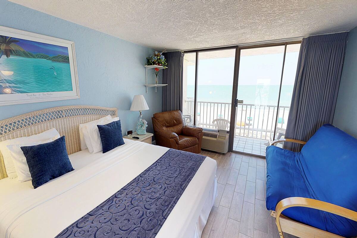 A hotel room with a large bed, a blue armchair, a balcony with a view of the sea, and a painting of a beach scene.
