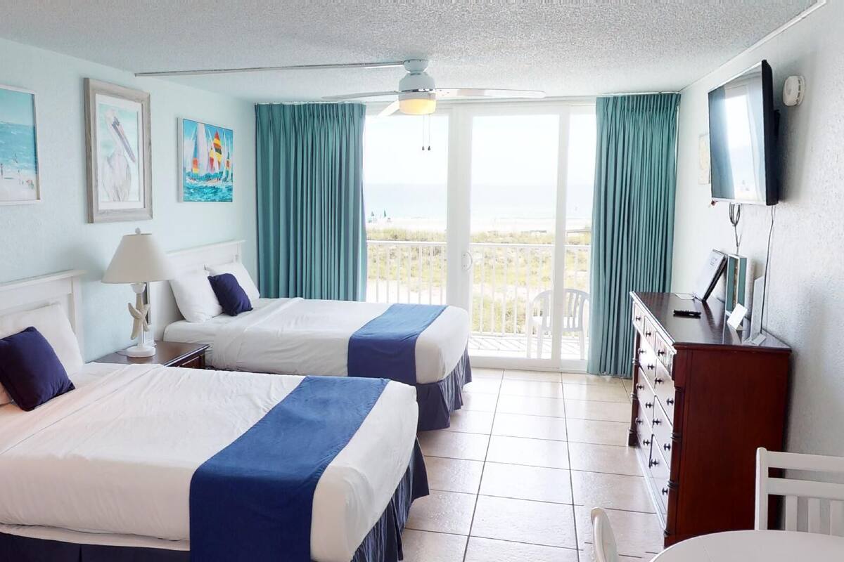 A hotel room with two beds, a desk, a chair, and a balcony with curtains.