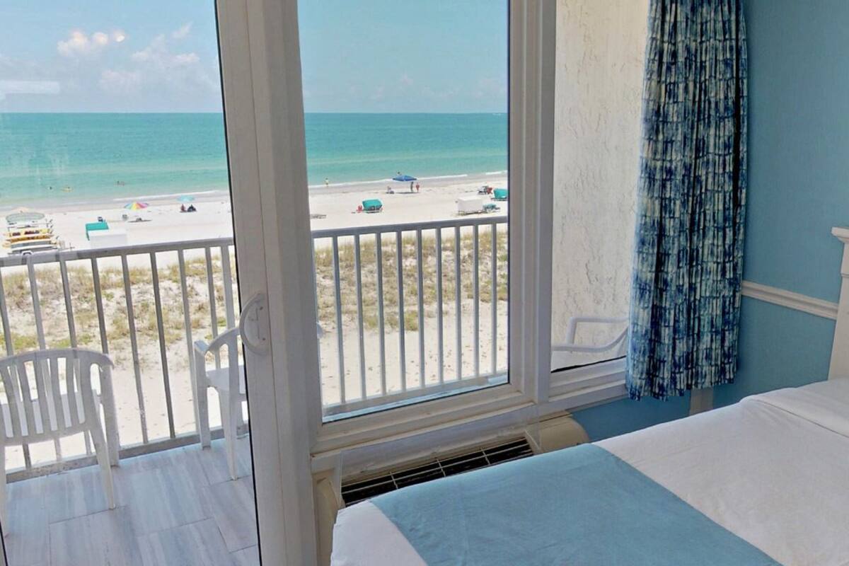 A hotel room with a large window overlooking a beach, a bed with white bedding, and a blue and white striped chair.