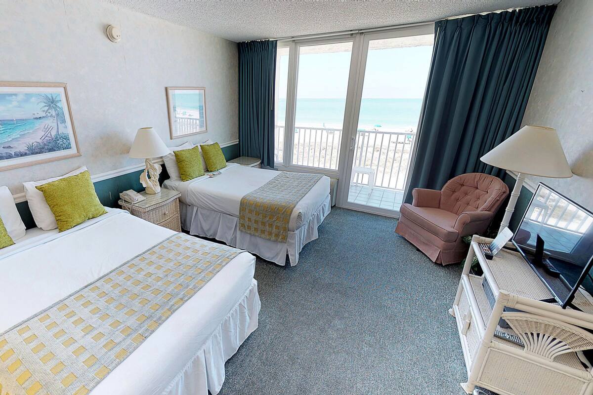 A hotel room with two beds, a chair, a TV, and a balcony with a view of the sea.