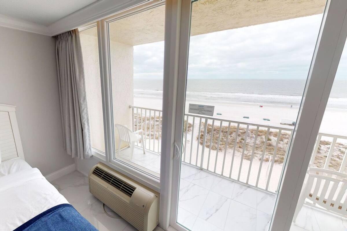 A hotel room with a large window overlooking a beach, a bed with white linens, and a ceiling fan.