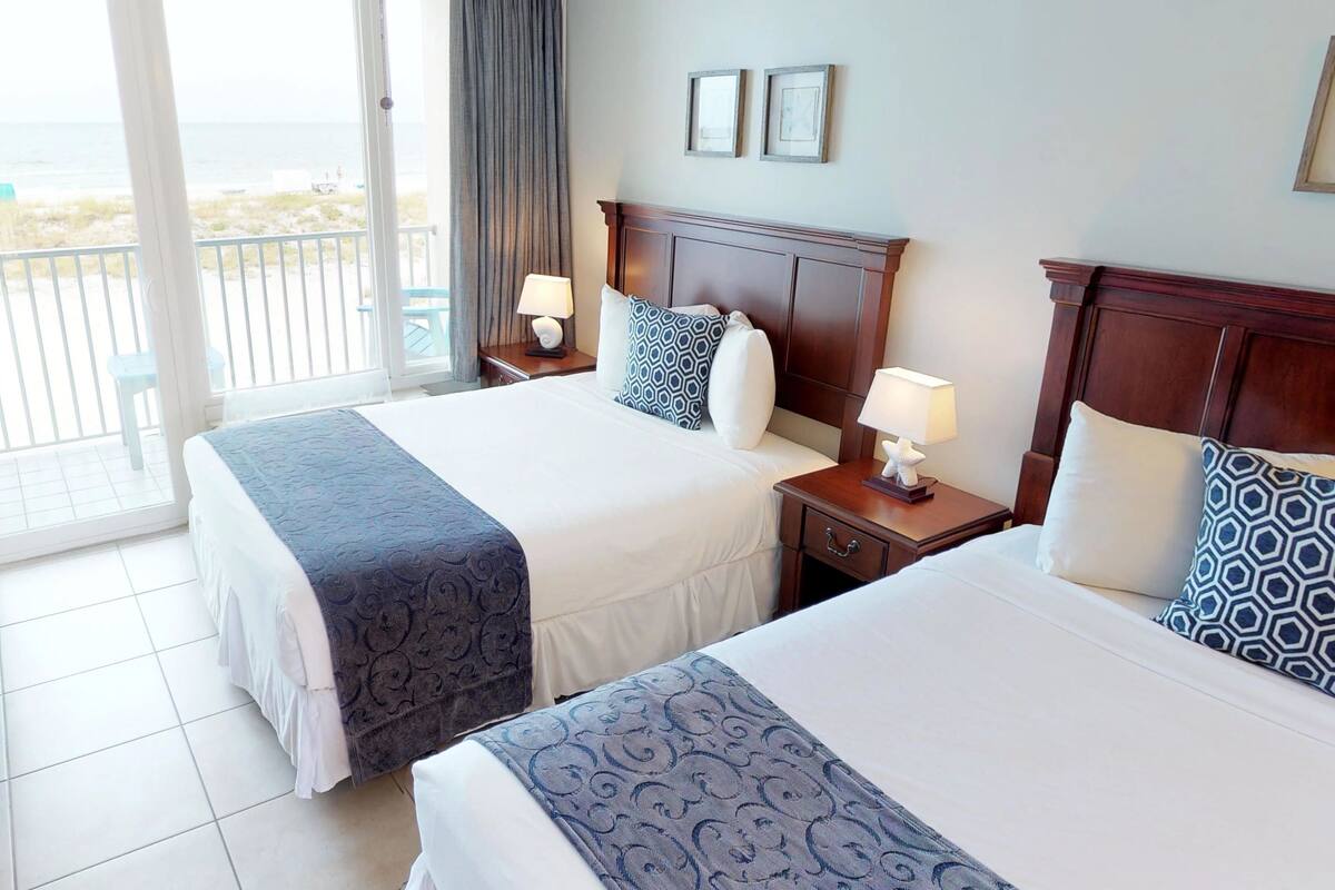 A hotel room with two beds, a chair, a nightstand with a lamp, and a view of the beach.