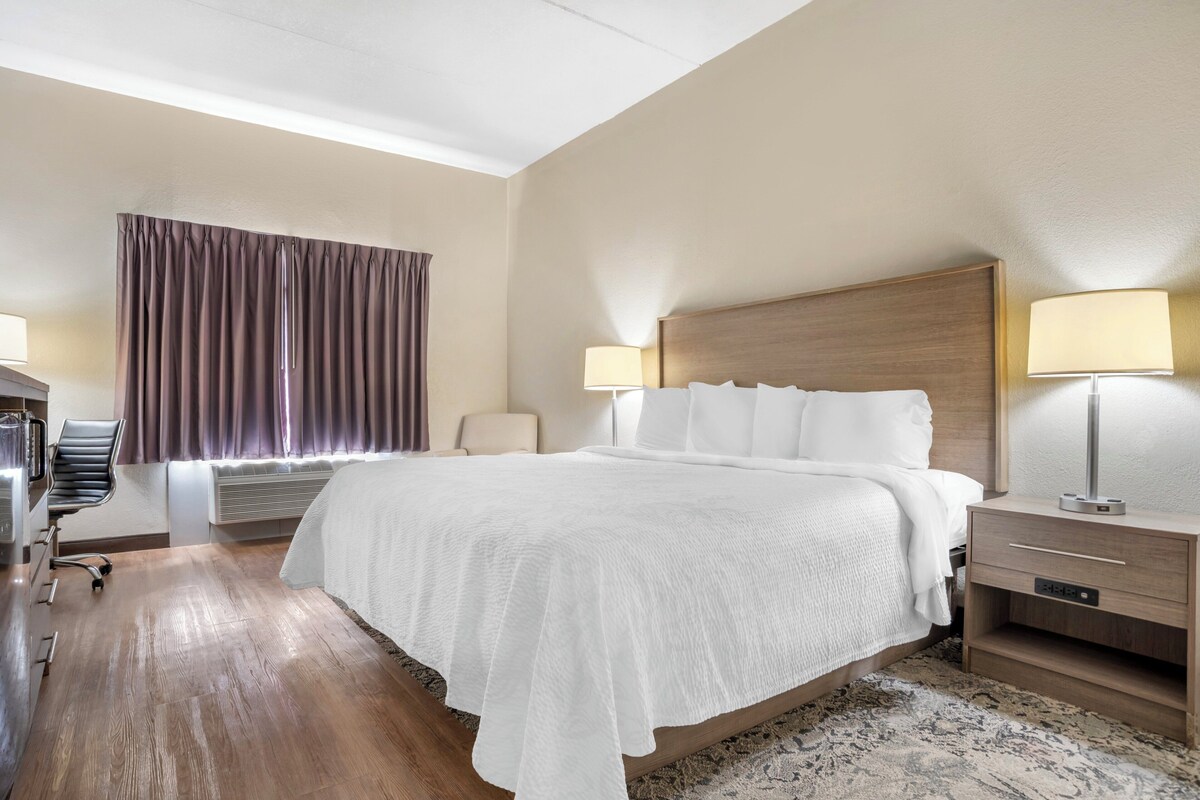 A hotel room with a large bed, two bedside tables with lamps, a chair, a desk, and a window with curtains.