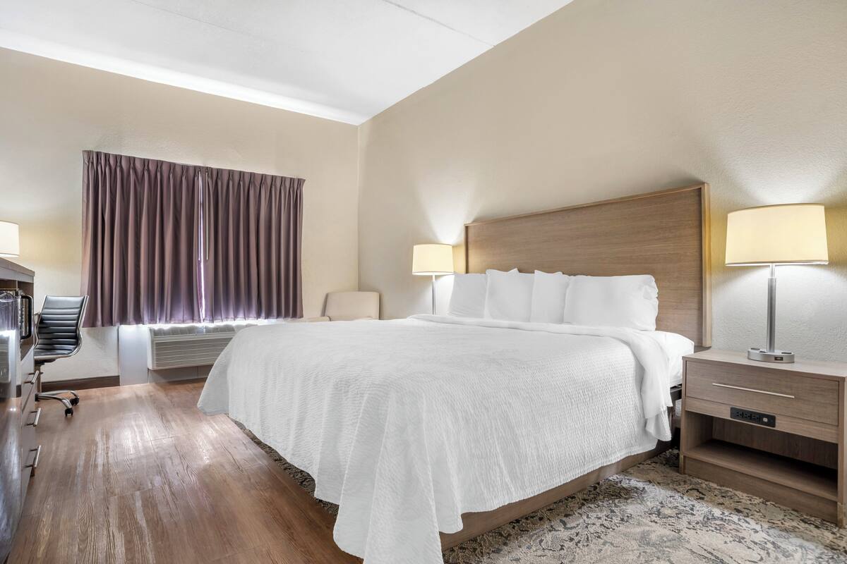 A hotel room with a large bed, two bedside tables with lamps, a chair, a desk, and a window with curtains.