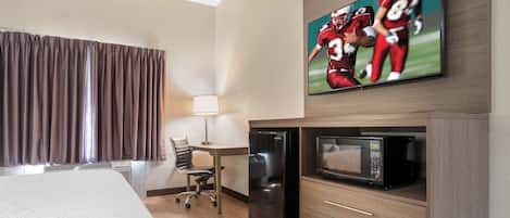 Habitación de hotel con cama, escritorio con refrigerador, microondas, silla y televisor montado en la pared.