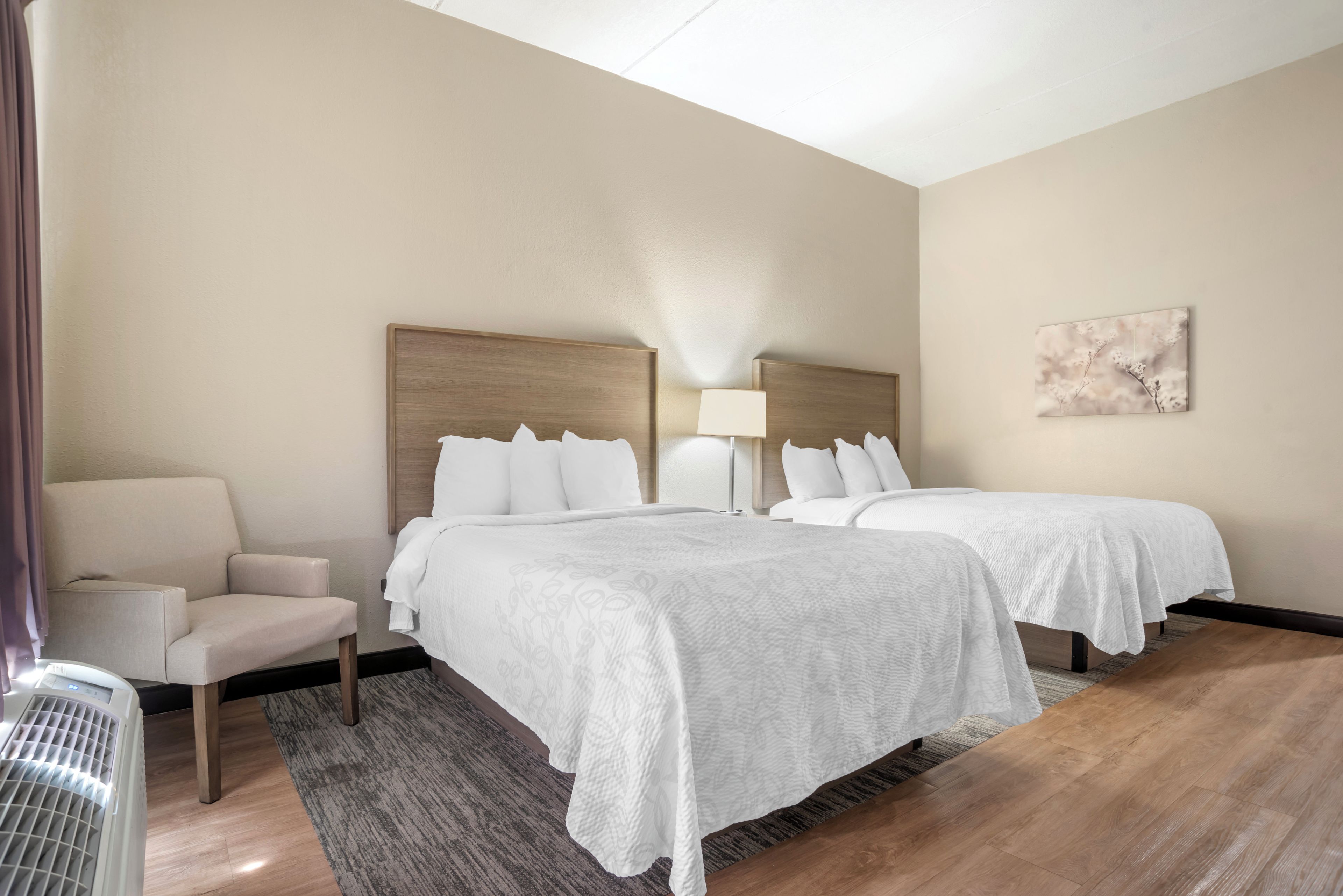 A hotel room with two beds, a chair, a lamp, and a painting on the wall.