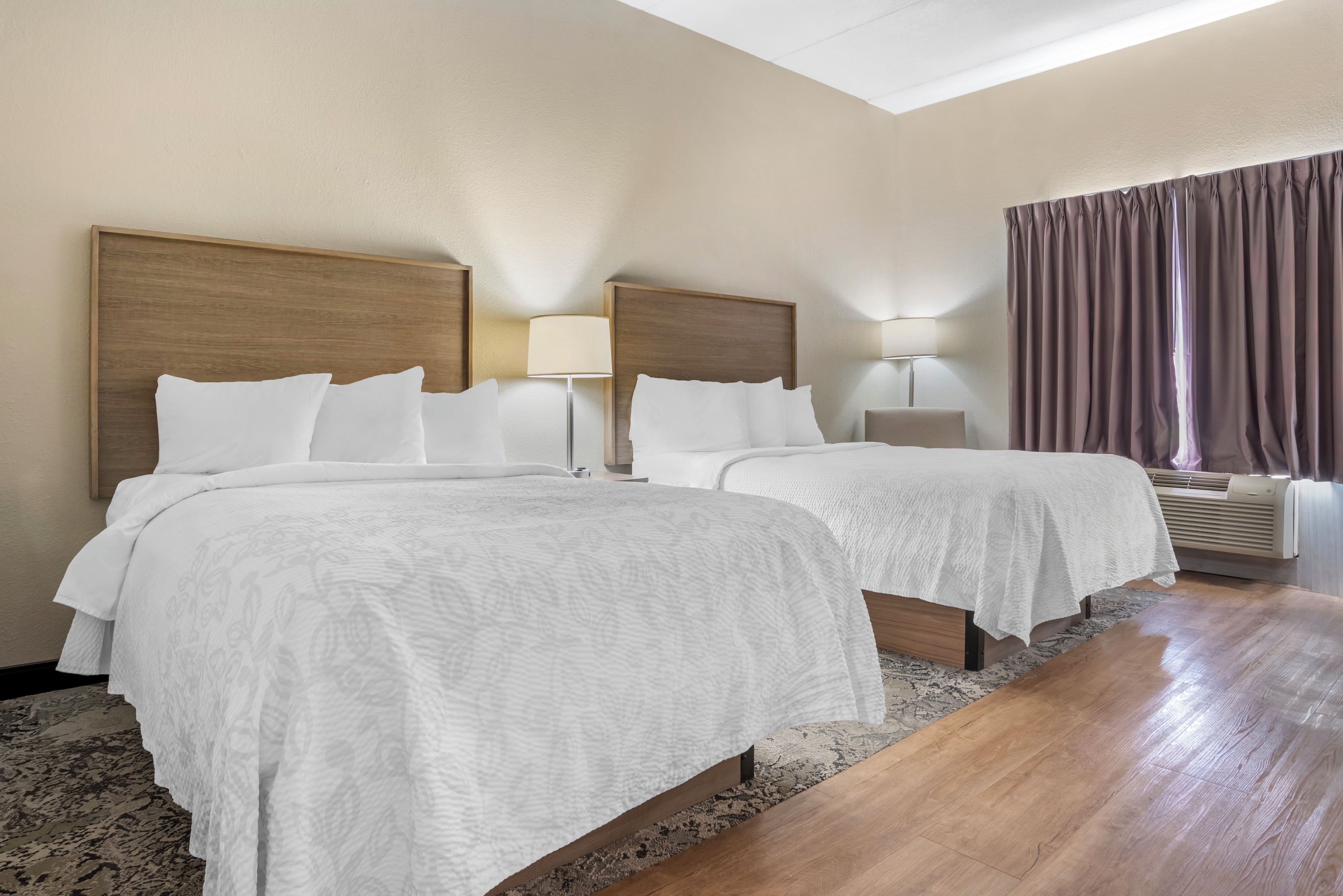 A hotel room with two beds, a large window with curtains, a standing lamp, and a ceiling light.