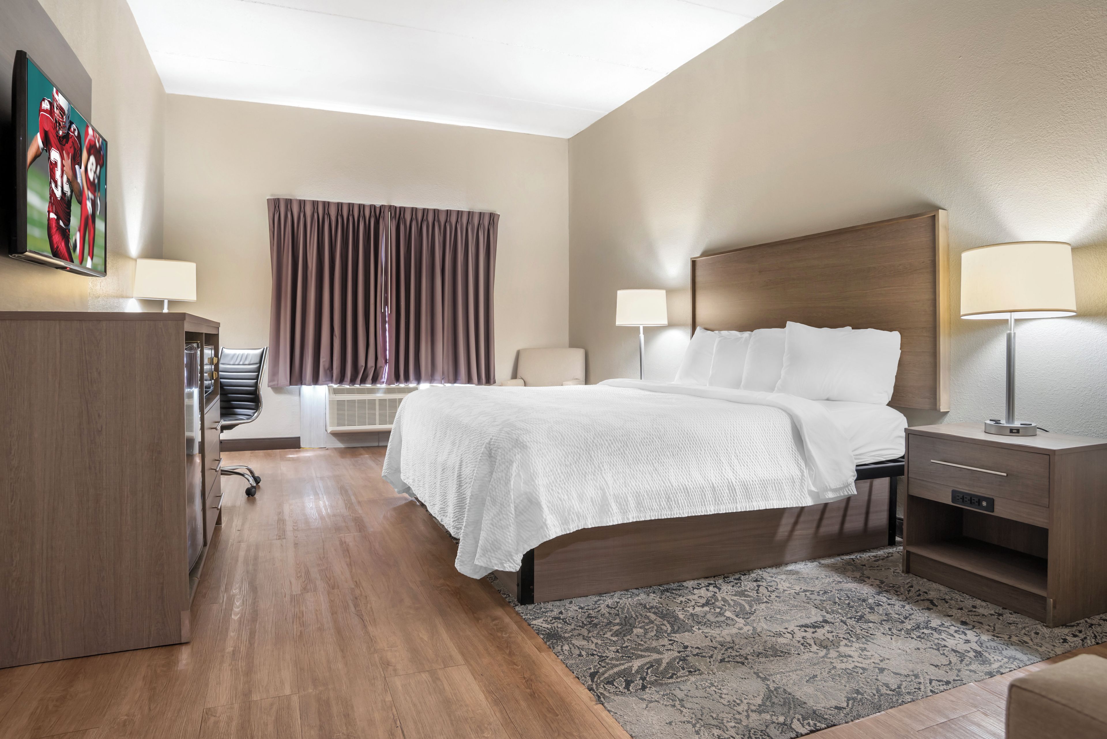 A hotel room with a bed, bedside tables, a desk, a chair, a window with curtains, and a wall-mounted TV.