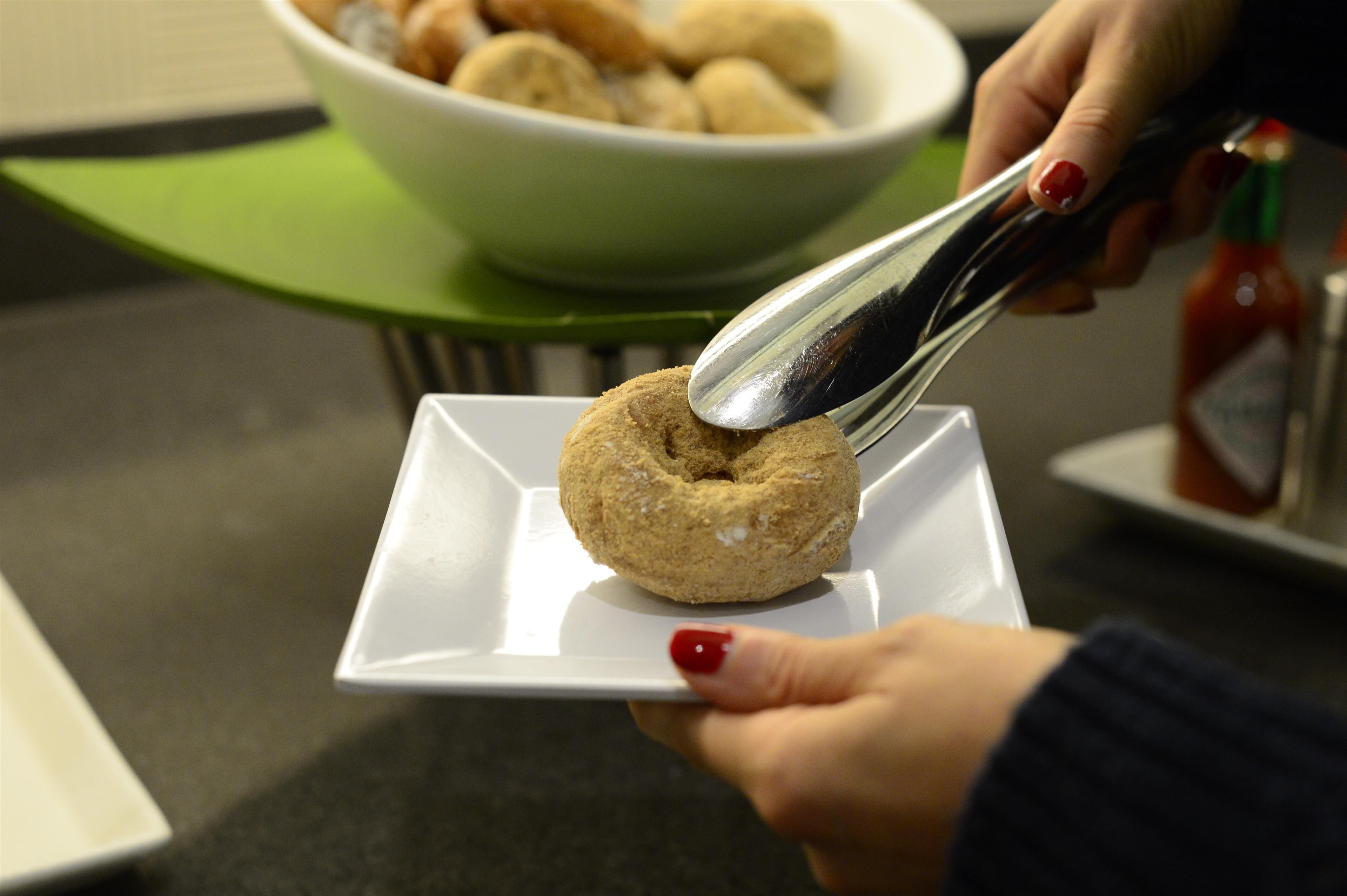 A hand holding a spoon over a square plate with a food item, a bowl of similar items in the background, and a bottle of sauce on a table.