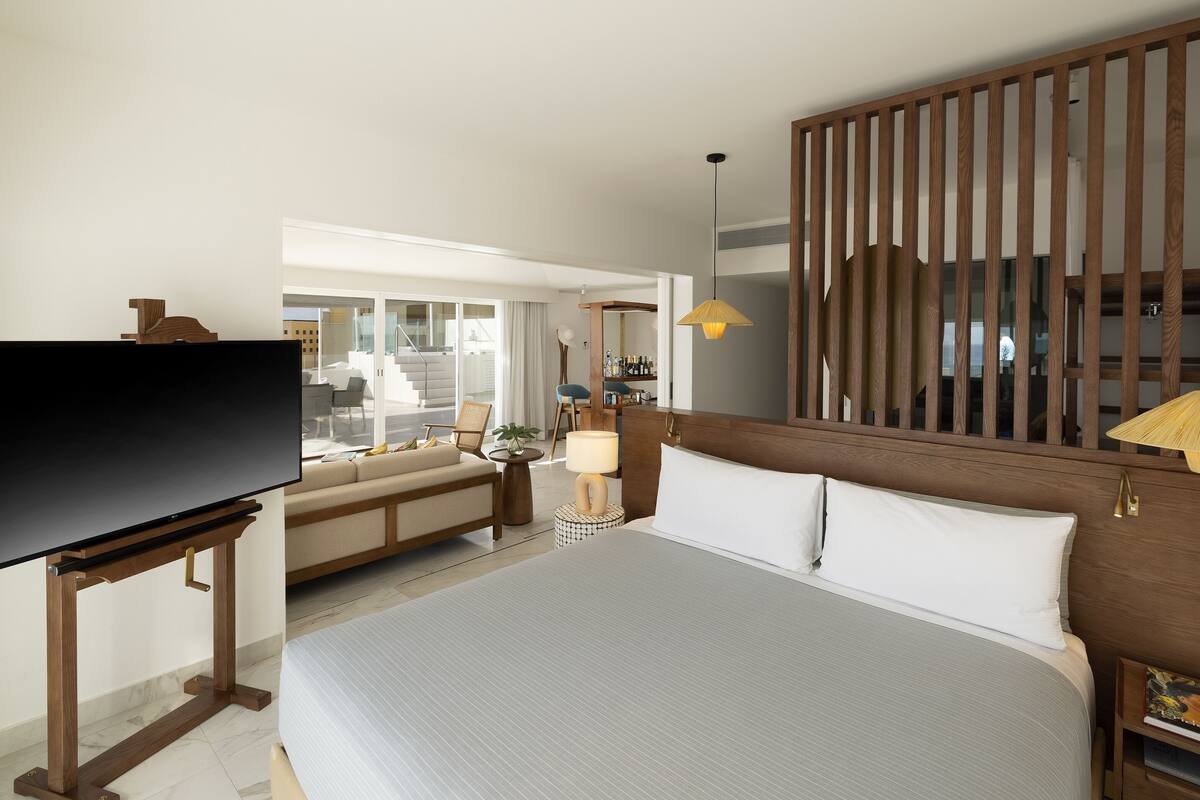 Una habitación de hotel moderna con una cama grande, un televisor sobre un mueble de madera, una mesita de noche con una lámpara y vistas al balcón exterior.
