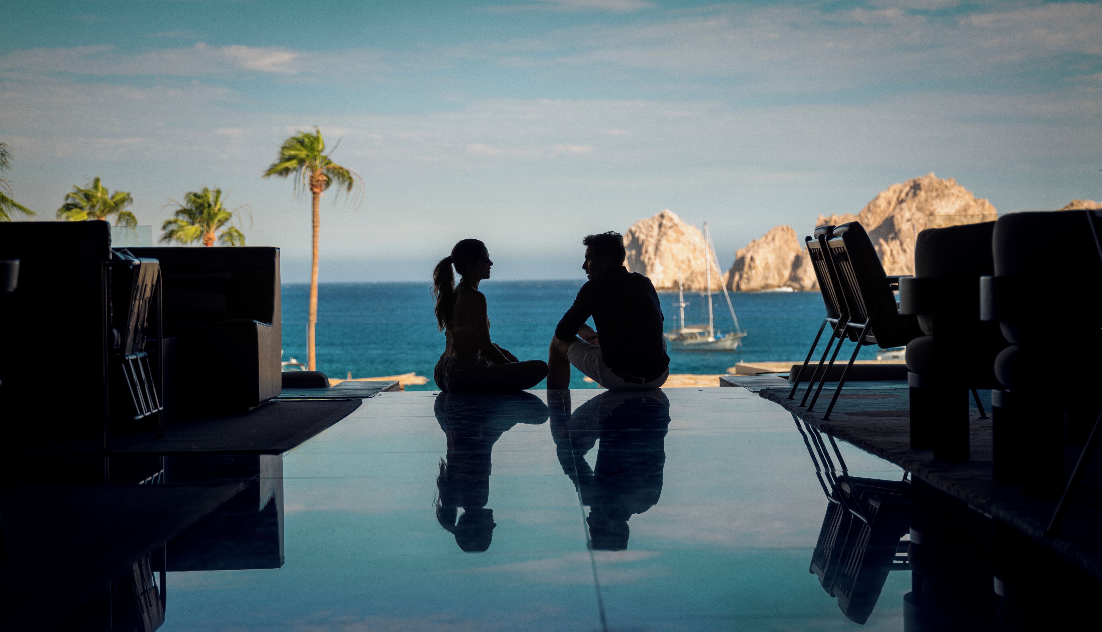 A couple sitting by a reflective pool with a view of the ocean and palm trees.
