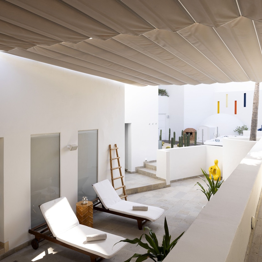 A sunlit outdoor area with white walls, a pool, lounge chairs, and a wooden ladder.