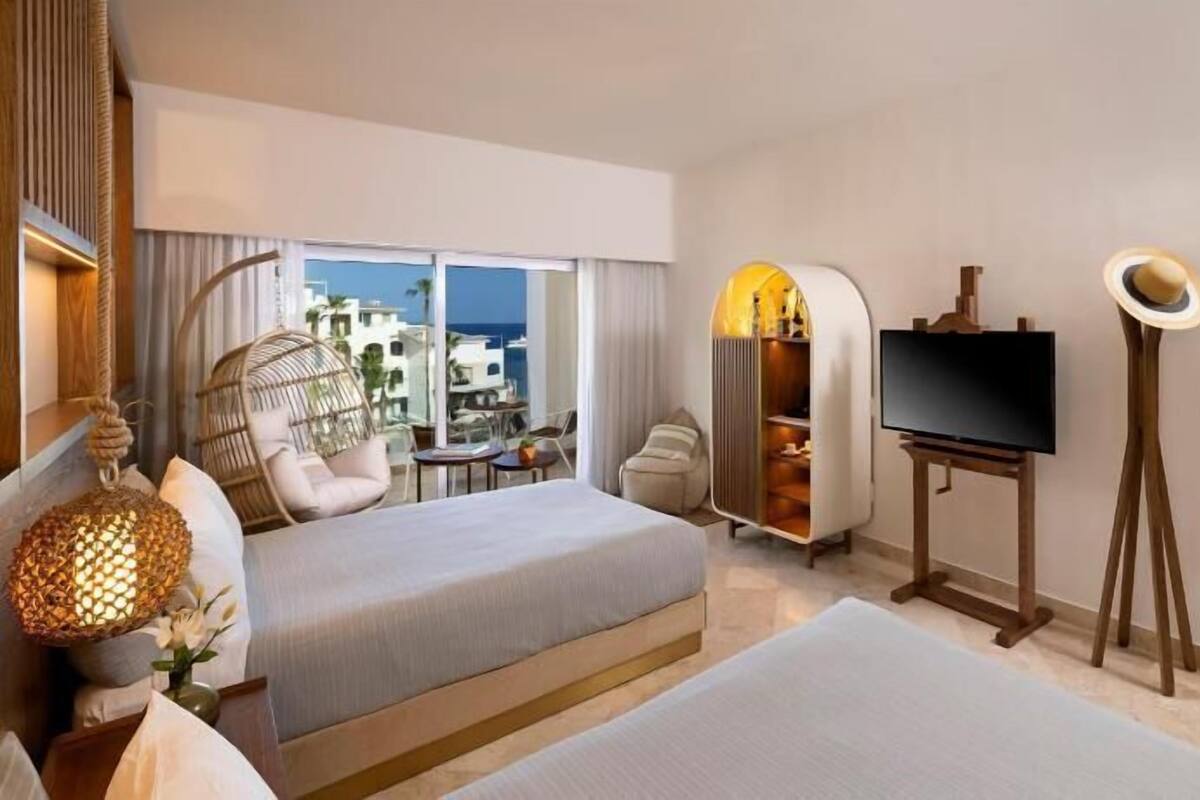 Habitación de hotel con cama, televisión, zona de estar con una silla y mesa, y vista al mar.