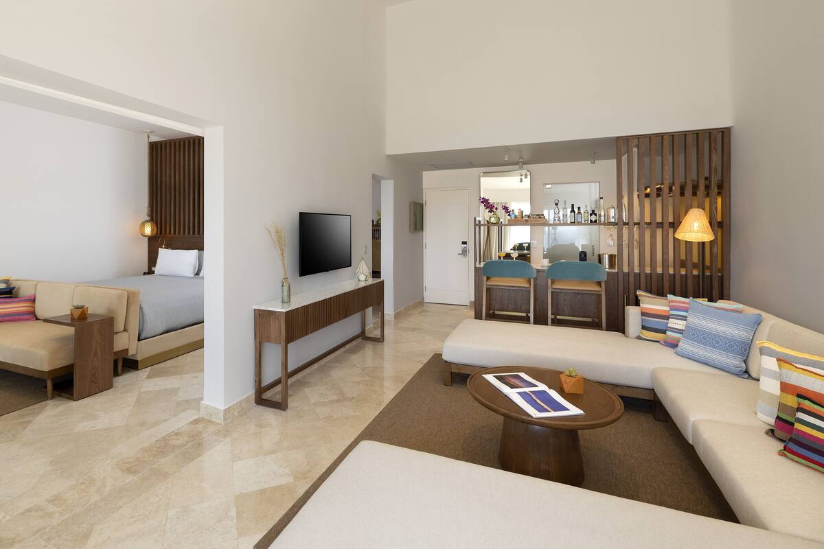 Una habitación de hotel moderna con sofá, cama, televisión y zona de comedor con mesa y sillas.
