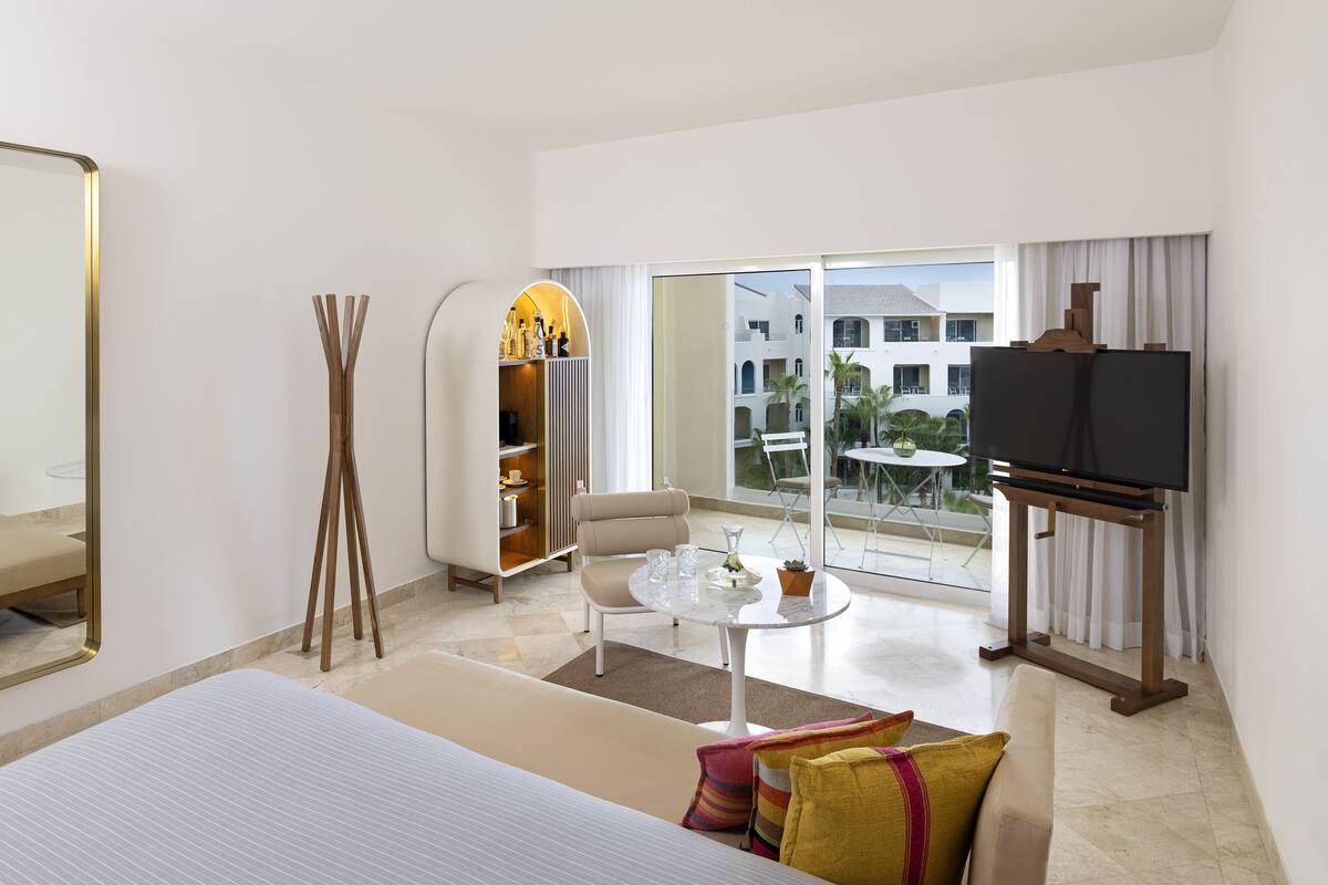 Una habitación de hotel moderna con una cama grande, una zona de estar con una mesa de centro, televisión y un balcón con vistas a edificios y zonas verdes.