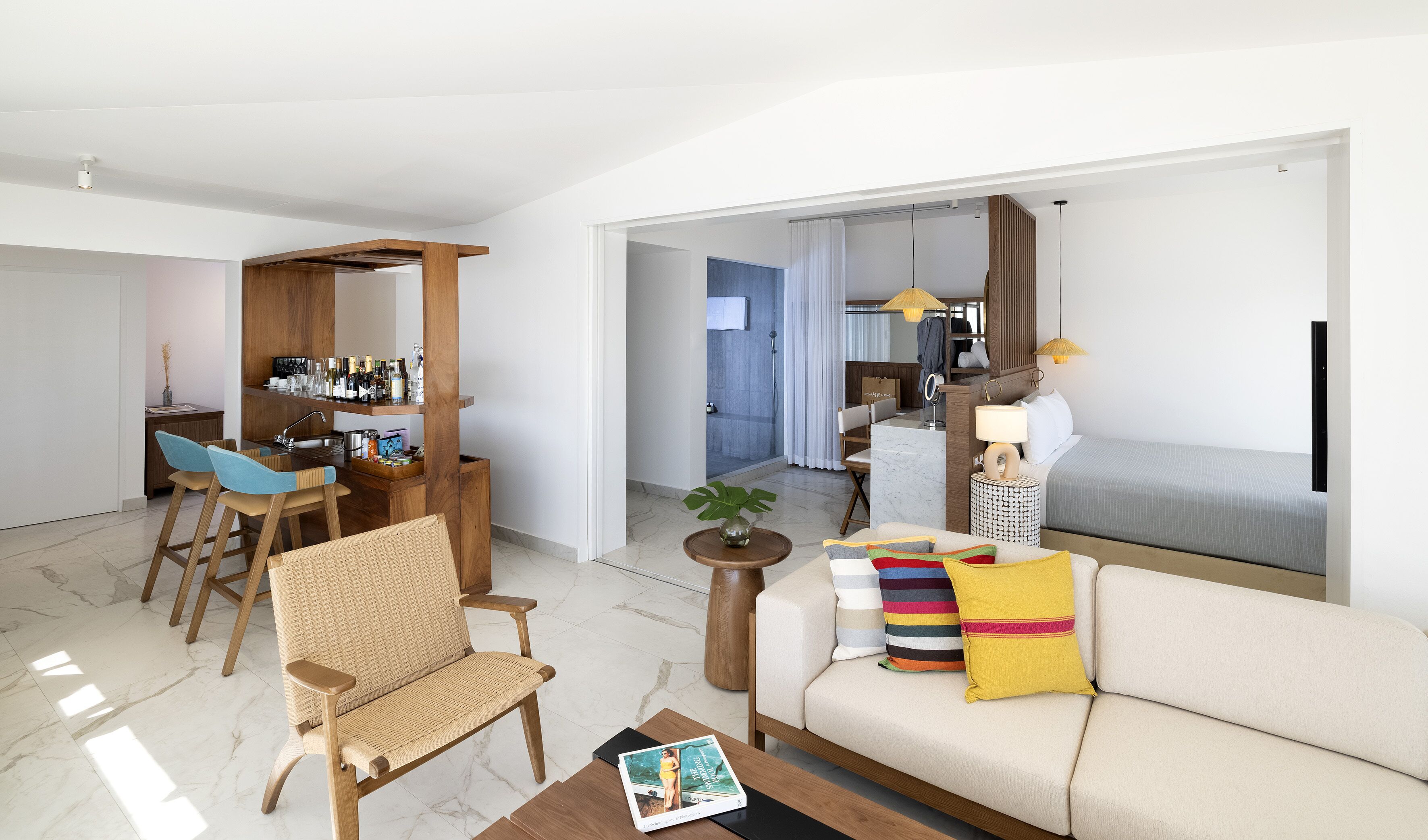 A modern hotel room with a sofa, a dining area with chairs, a bar with bottles, a shower area, and a bed.