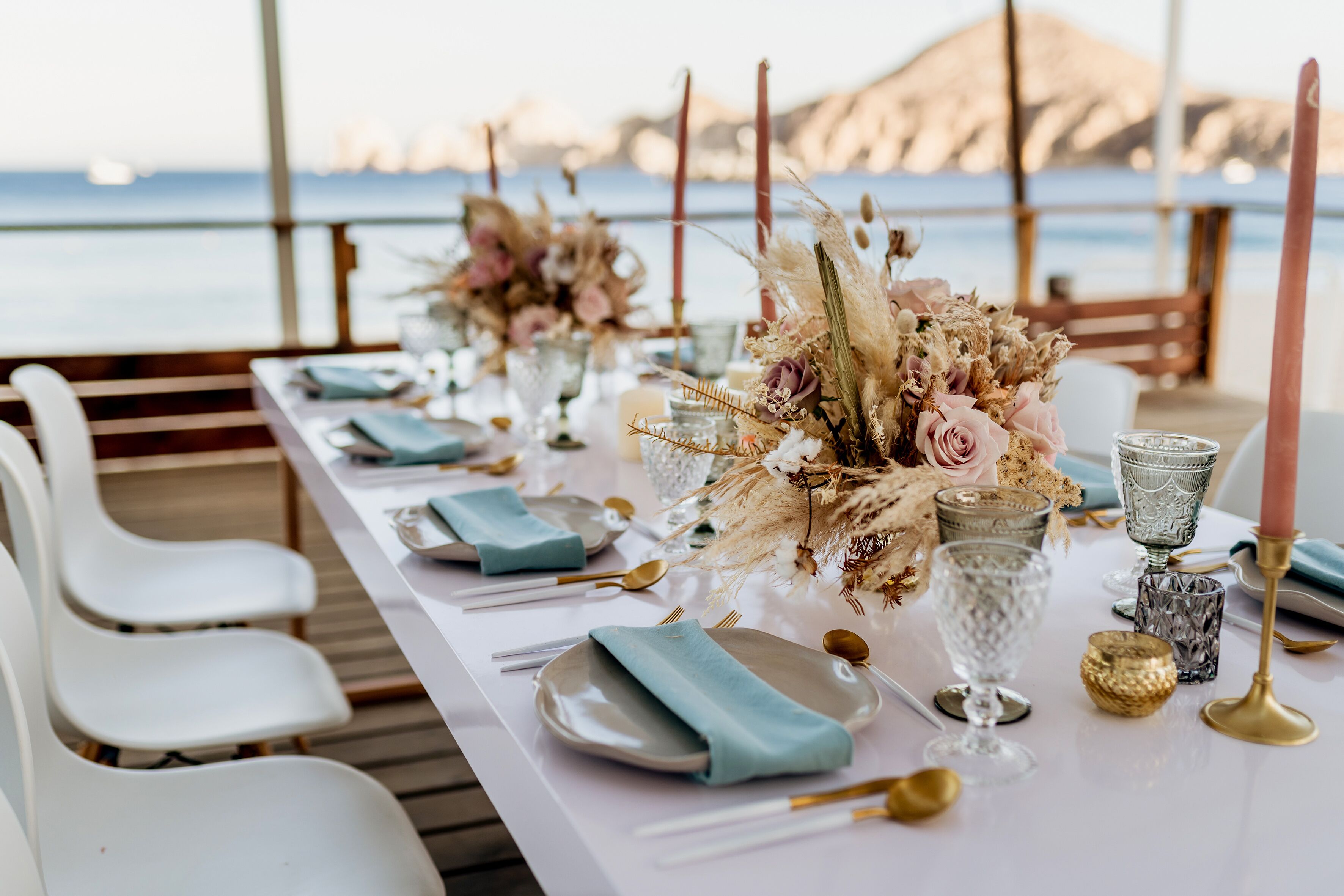 Una mesa dispuesta con platos, vasos y cubiertos, adornada con flores y velas, con vista a un cuerpo de agua.